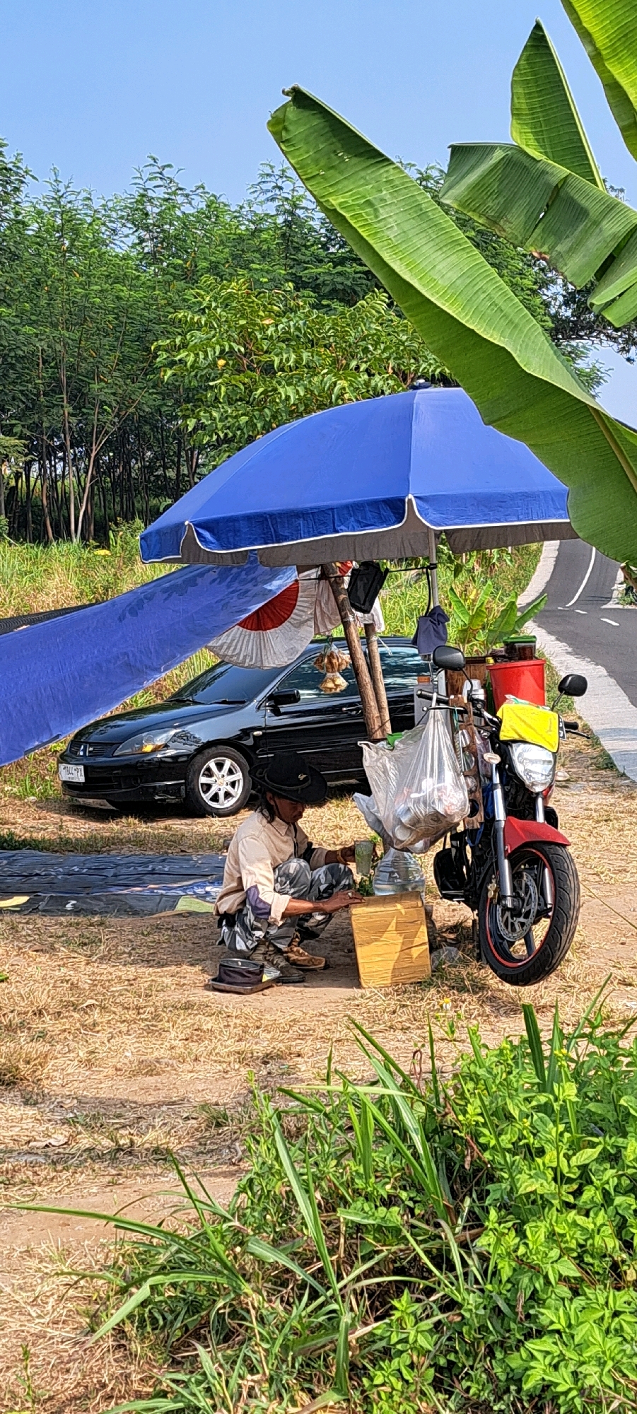 In Sekarjoho, Indonesia, a person wearing a light-colored long-sleeved shirt, camouflage pants, a dark hat, and sandals squats beside a wooden crate. They are holding a green cup and appear to be preparing something on the crate, which is placed on dry, brown ground. Behind the person, a black car is parked. A large blue umbrella, supported by wooden poles, is set up, casting shade. Hanging from the umbrella poles are a few items, including a clear plastic bag filled with objects. To the right of the umbrella, a red bucket and a red motorcycle are visible. The motorcycle has a yellow cover on its front and bags hanging from its handlebars. In the foreground, green grass and plants grow. In the background, a paved road curves upwards, lined with trees and foliage, with a clear blue sky above. Banana leaves are prominent in the upper portion of the image.