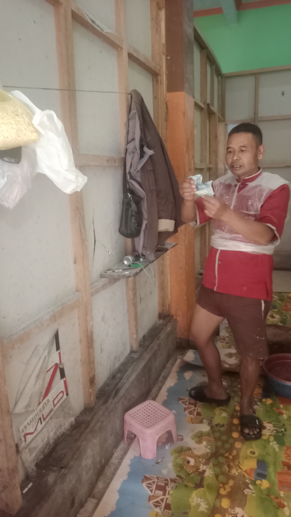 The image shows a man in a red and white shirt and brown shorts holding several Indonesian Rupiah banknotes. He appears to be standing in a room with unfinished wooden walls and a light-colored, possibly cracked, wall surface. Hanging on the wall are a brown jacket and a black bag. A small shelf below the hanging items holds a can and other small objects. In the foreground, a pink plastic stool is visible on a floor with a colorful mat depicting cartoon houses and nature. The man is holding money in his hands and looking towards it. The environment suggests an indoor space, possibly a home or a workshop, with visible signs of construction or disrepair.
