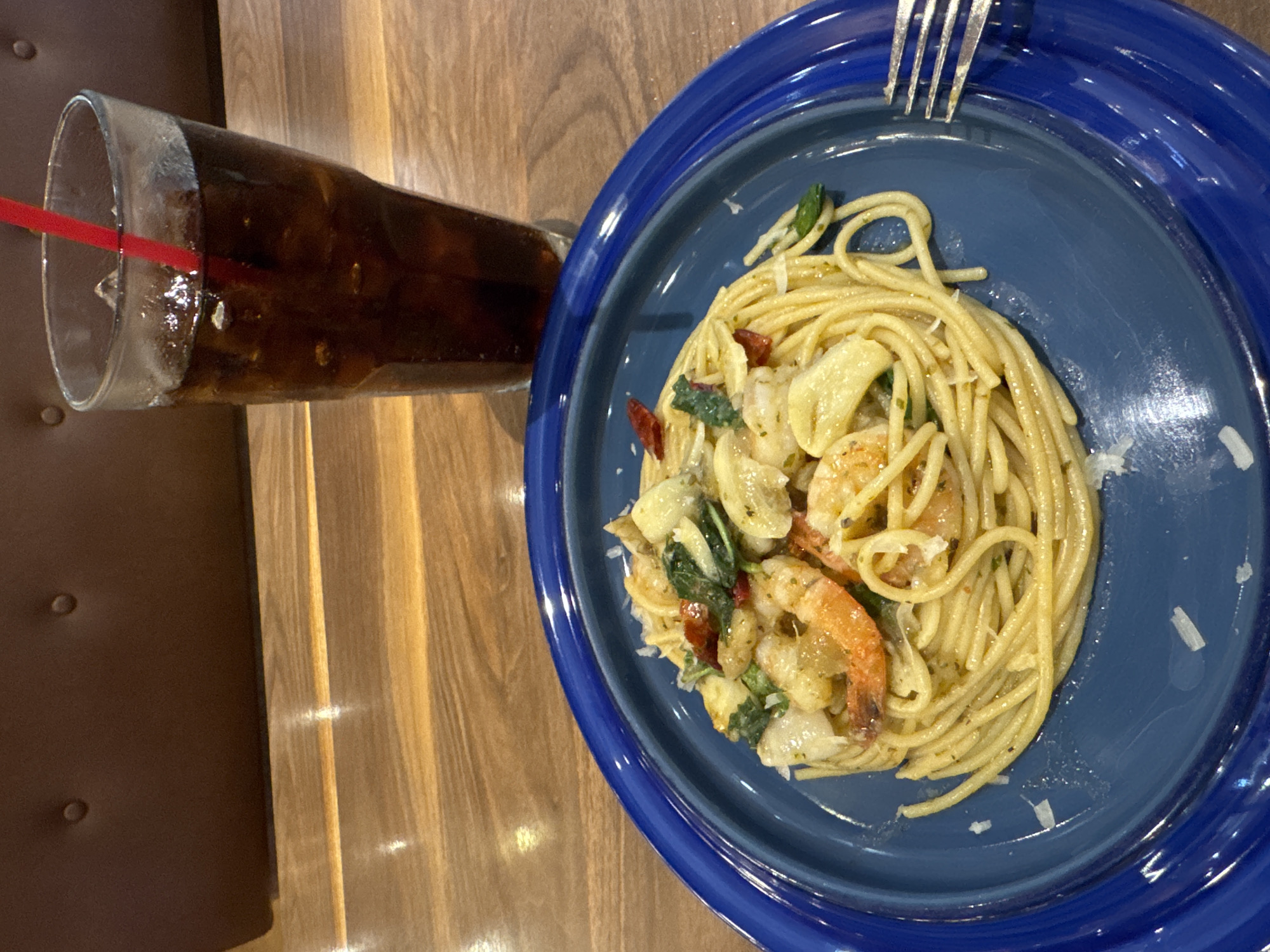 A close-up, eye-level shot shows a blue plate filled with spaghetti and shrimp. The pasta is coiled in the center of the plate, topped with several pink shrimp, chopped green herbs, and thin slices of red chili peppers. A sprinkle of grated white cheese is visible on the pasta. To the right of the plate, a portion of a silver fork rests on the blue rim. Behind the plate, a tall glass filled with a dark carbonated beverage and ice cubes, with a red straw, stands on a wooden table. In the background, a portion of a dark brown, tufted upholstered booth is visible. The image is taken in a restaurant setting. There is no information in the image to suggest a specific location in the city of Wichit, Thailand.