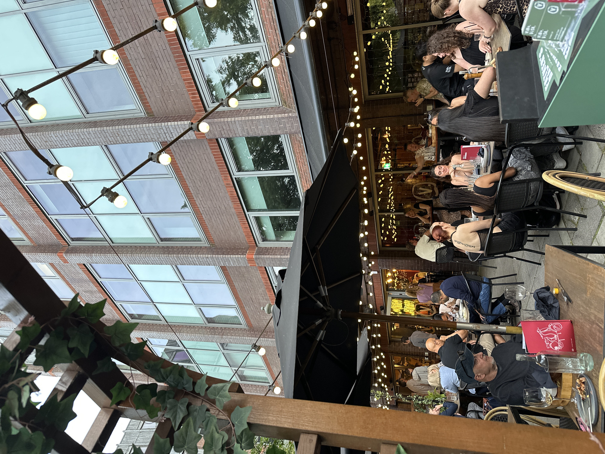 An outdoor patio dining area is depicted, with patrons seated at tables. A brick building with large windows forms the backdrop, and string lights are draped overhead. The scene appears to be in the evening, with ambient lighting from the string lights and interior of the building. One man in the foreground wears a black t-shirt and baseball cap, looking towards the right of the frame. He is seated at a wooden table with wine glasses, a bottle of wine, and a red menu with a scooter illustration. Several other tables with diners are visible, and some are partially obscured by an umbrella. The atmosphere suggests a social gathering or meal at a restaurant.