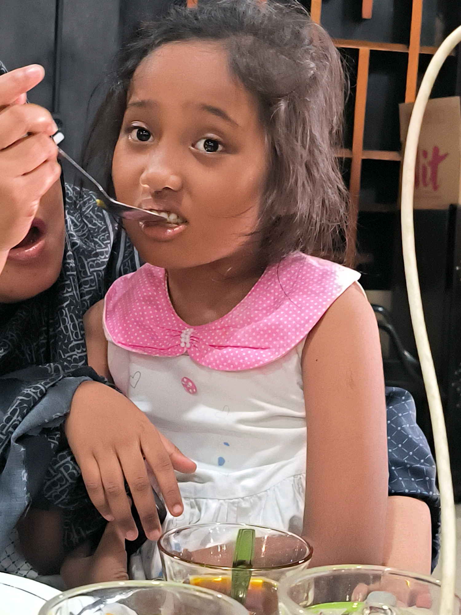 A young girl with dark hair and brown eyes is being fed with a spoon by someone off-camera. She is wearing a white dress with a pink collar adorned with white polka dots and small pearl-like beads. The collar has a pink ribbon tied into a bow. Her left arm is resting on the arm of an unseen person, whose dark blue garment with a white pattern is visible.
The girl is looking towards the camera with a slightly surprised expression, her mouth open as the spoon with food is being offered to her. In the foreground, two glass bowls are visible, one containing a yellowish liquid with a green element, and another partially obscured by the first.
The background is dark and out of focus, showing what appears to be a wooden grate-like structure and a glimpse of a brown paper bag with pink lettering. The lighting suggests an indoor setting, possibly during the day. The overall scene captures a moment of a child being fed, with a warm and intimate feel. The location context is Banda Aceh, Indonesia, though no specific visual cues within the image definitively confirm this. No visible text is legible except for the partial pink lettering on the paper bag, which is too indistinct to read.