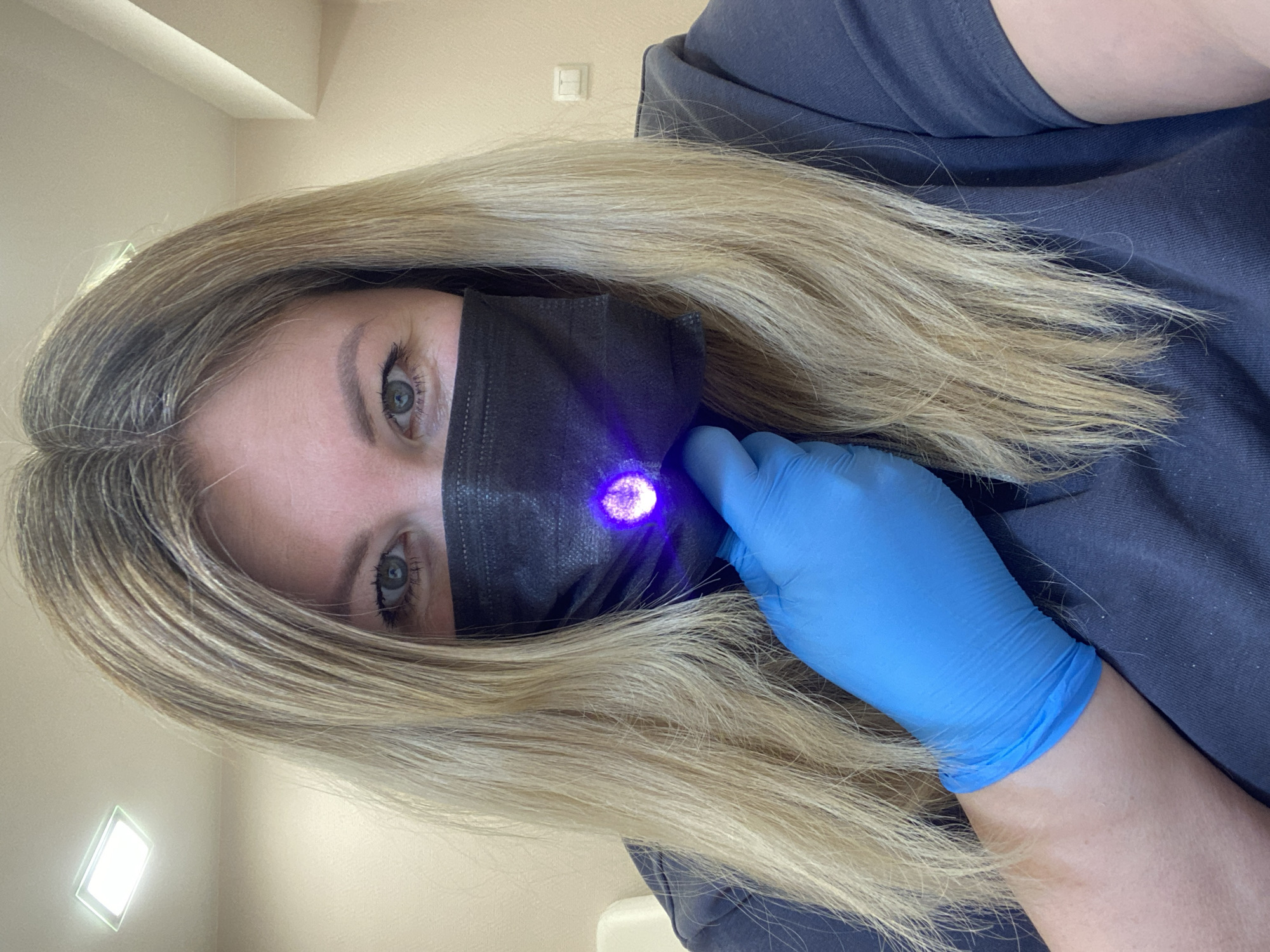 A person with light blond hair is wearing a dark gray shirt, a black disposable face mask, and a blue latex glove. The person is holding a blue light near the center of the mask. The location appears to be indoors, with a light fixture visible in the upper left corner of the image. There is no information to suggest the image was taken in Chaikovsky, Russia.
