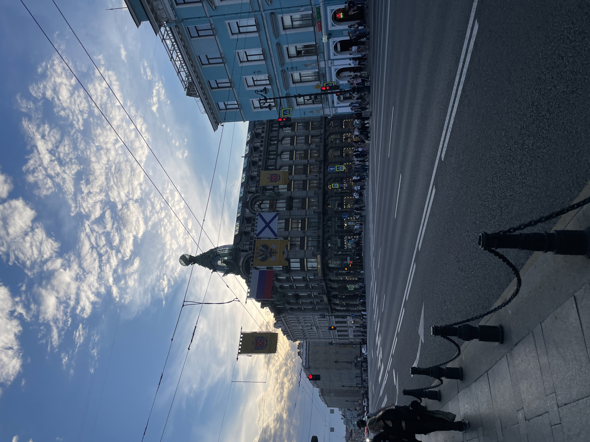 An upward-angled, daytime photograph captures a vibrant street scene in Saint Petersburg, Russia. The image focuses on a grand, multi-story building on the left, identified as Dom Knigi (Singer House) by the visible text on its facade, featuring an ornate domed corner tower. Several historical and national flags adorn this building, including the St. Andrew's flag (white with blue saltire), a yellow flag with a double-headed eagle (Imperial Russian coat of arms), a Russian tricolor flag, and another yellow flag with a coat of arms.
To the right and slightly behind the main building is another distinctive light blue building. Its facade displays Cyrillic text above an entrance, partially visible as "АКАДЕМИЯ..." (Academy...). The wide asphalt road in the foreground and mid-ground features clear white lane markings, with two red traffic lights indicating a stop. A sidewalk lines the right side of the road, marked by decorative dark chain-link posts.
Numerous electrical wires crisscross the bright blue sky, which is dotted with scattered white, fluffy clouds. The low angle of the sun, likely late afternoon or early evening, casts a soft, warm glow on the clouds and highlights parts of the buildings. People are visible strolling on the sidewalks, and some are waiting near the traffic lights. The scene portrays typical urban activity, with the prominent historical architecture and flags providing strong location cues for Saint Petersburg.