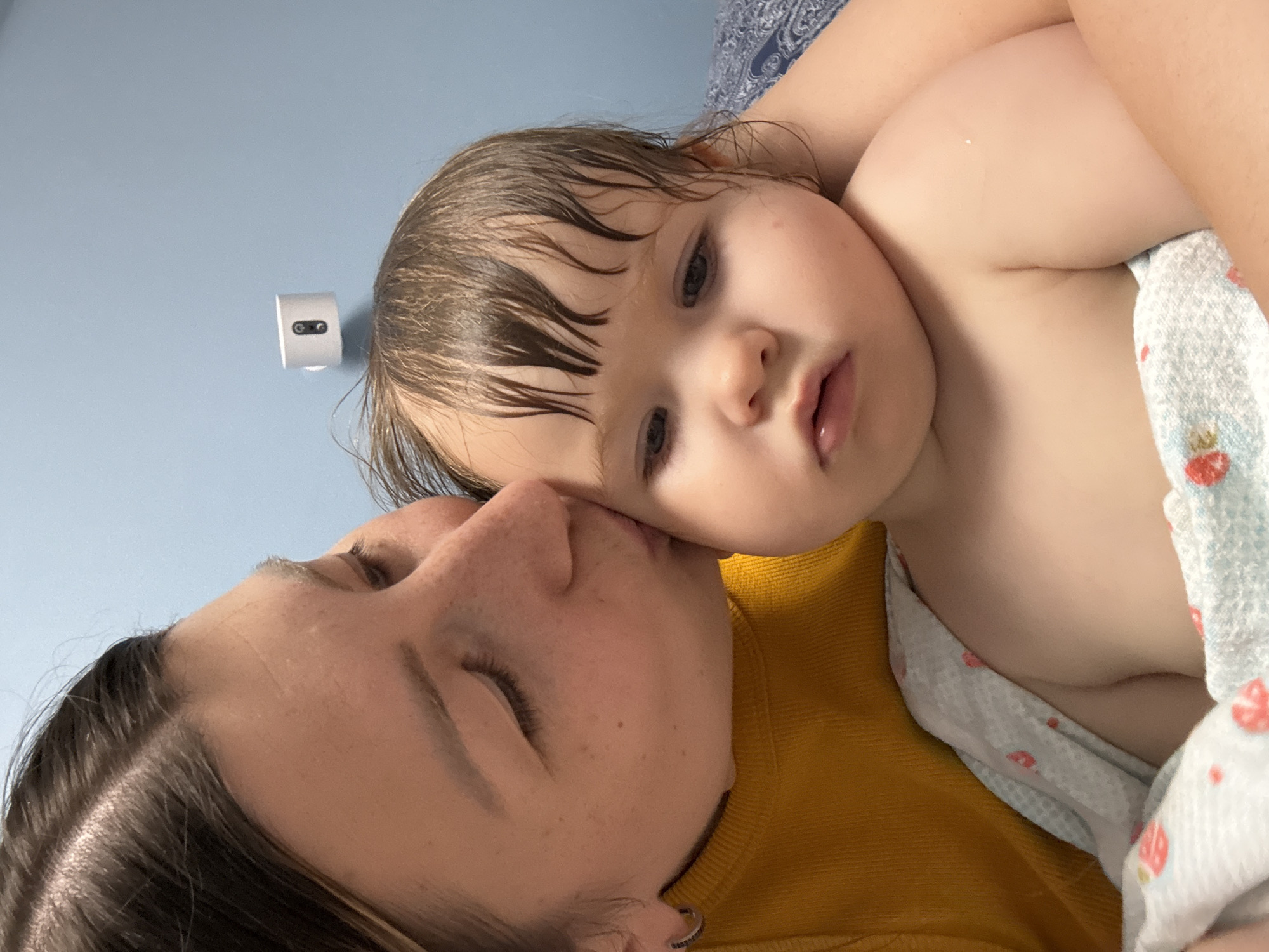A close-up, head-and-shoulders shot captures a woman kissing a baby. The woman, on the left, wears a yellow top and has light brown hair. Her eyes are closed, and she is kissing the baby on the cheek. The baby, on the right, is looking down with a neutral expression. The baby has light brown hair, which appears damp, and is wrapped in a light blue and white patterned blanket. The background consists of a light blue wall with a small white object mounted on it, resembling a motion sensor or camera. The overall lighting is bright, and the focus is on the subjects. The image does not contain any visual information that can be used to determine its location within Moscow, Russia.