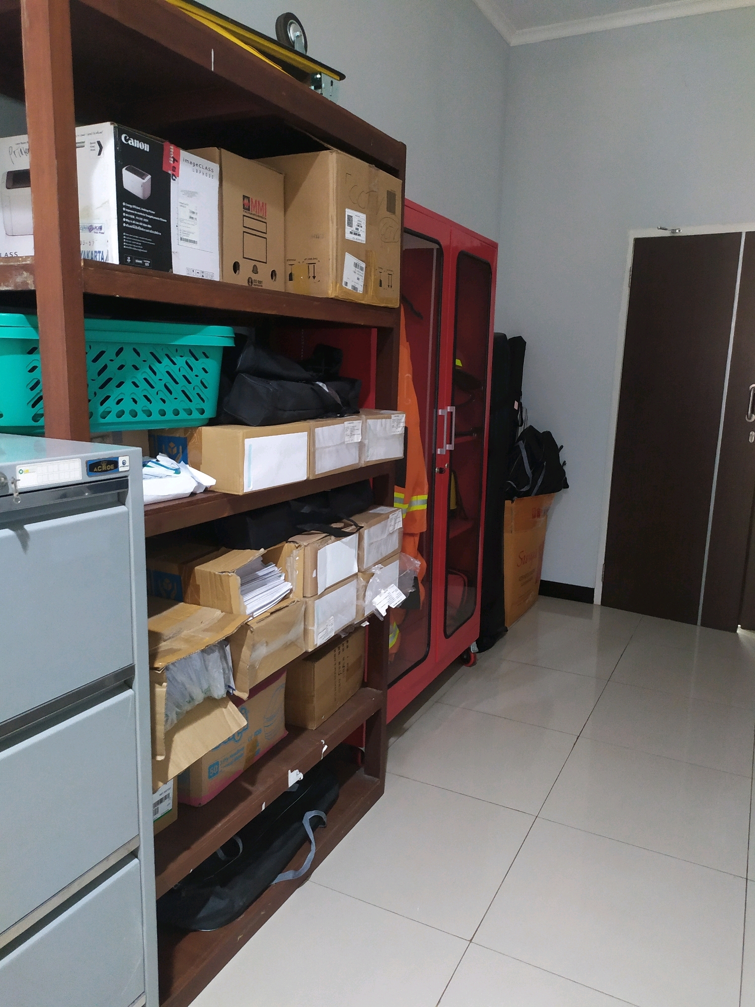 The image depicts an indoor storage area, likely a utility room or office supply room, in Wates, Indonesia. Dominating the scene is a tall, wooden shelving unit filled with various boxes and containers. On the top shelf, several cardboard boxes are visible, including one labeled "Canon". Below that, two teal plastic baskets are stacked. The middle shelves contain an assortment of cardboard boxes, some open and revealing their contents, such as papers and other packaged items. A light grey metal filing cabinet with multiple drawers is positioned to the left of the shelves. To the right of the shelving unit is a bright red metal cabinet with glass doors, containing an orange safety vest. Several black bags and a long, dark case are placed on the floor next to the red cabinet. The room has light blue walls and a tiled floor. A closed dark brown door is visible in the background on the right. The overall impression is one of organized, albeit somewhat cluttered, storage.