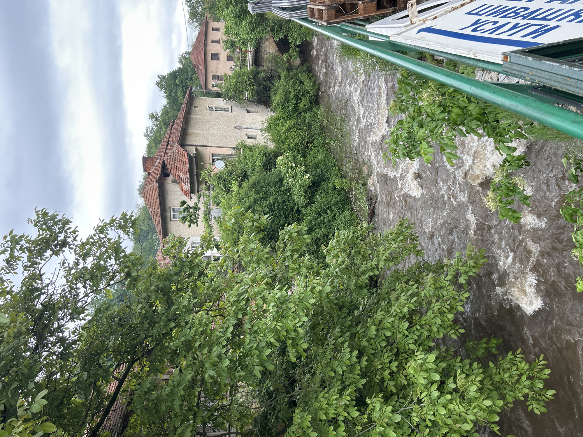 A landscape photograph taken on an overcast day shows a swift-flowing river coursing through an urban environment in Unknown. The river is muddy brown and foaming, indicating a high water level. On the left side of the river, there are multiple buildings with beige facades and red-tiled roofs partially obscured by lush green trees and foliage. A foreground tree with green leaves partially obscures the view, adding depth to the image. A bridge, with green railings and a white sign bearing blue lettering, crosses the river. The sign is oriented perpendicular to the camera, partially obscuring the view of the river and buildings.