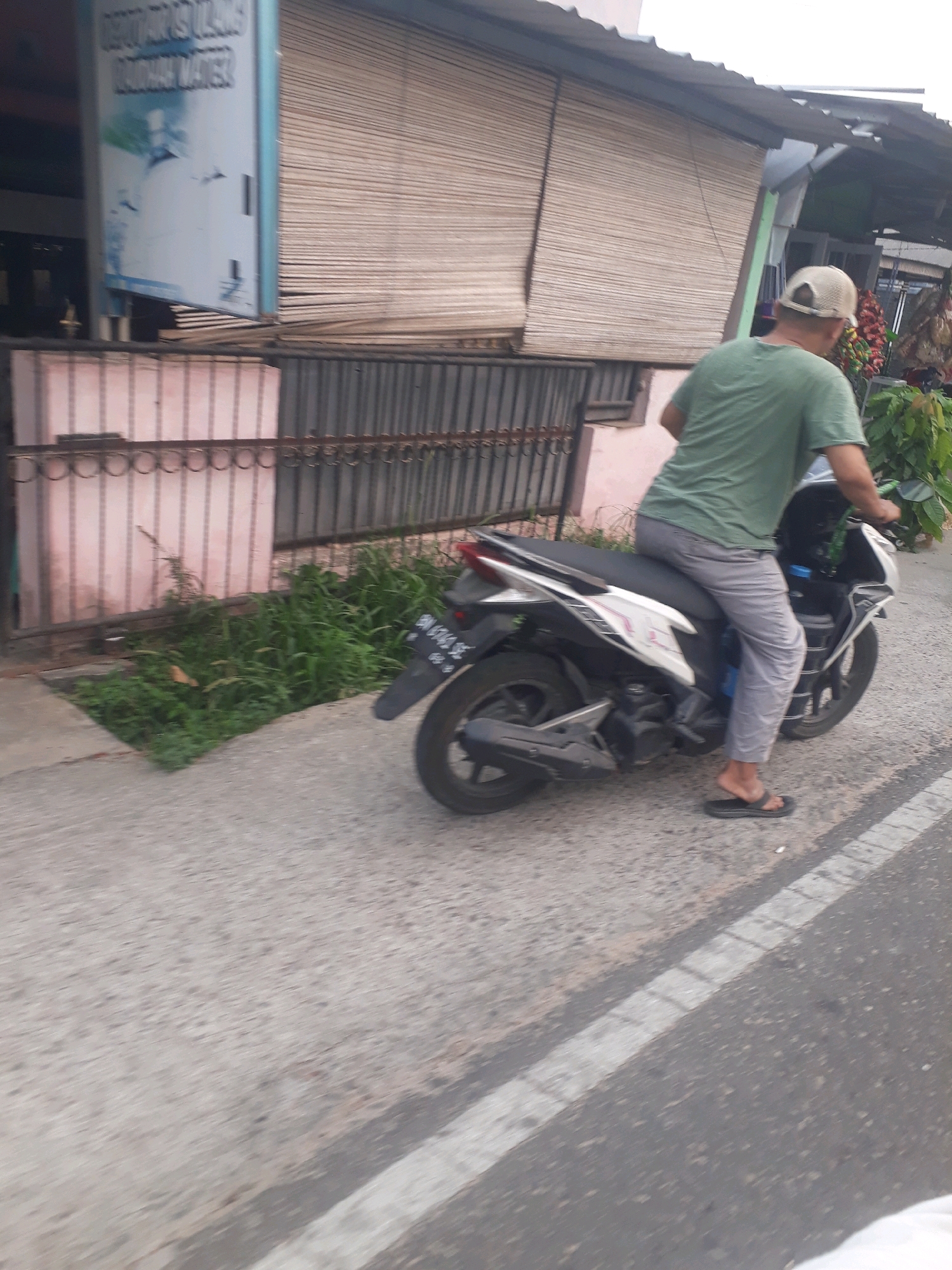 A person wearing a green t-shirt and gray pants is riding a white and black scooter on a paved road. The rider is wearing a light-colored baseball cap and black flip-flops. The scooter has a license plate that reads "BN 6343 SE". To the left of the road, there is a sidewalk with some overgrown green grass and a pink metal fence. Behind the fence, there is a building with a beige roll-up door. The scene appears to be in a semi-urban area, possibly a residential street or a commercial area, in Pangkalpinang, Indonesia, given the license plate format. The lighting suggests it is daytime.