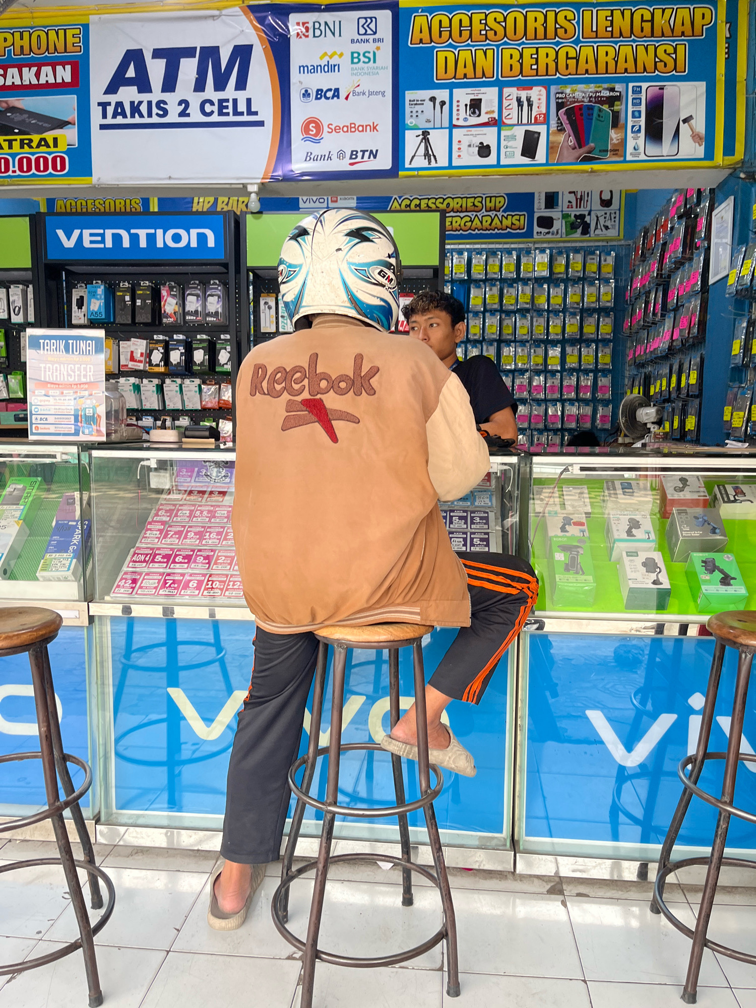 The image shows an outdoor stall in Yogyakarta, Indonesia, selling mobile phone accessories. A person wearing a helmet and a brown jacket with a Reebok logo sits on a stool, facing away from the camera. Another person, presumably an employee, is visible behind the counter, looking towards the left. The stall displays a variety of mobile phone accessories, including phone cases, chargers, and headphones. There are also signs for "ATM TAKIS 2 CELL" and "ACCESORIS LENGKAP DAN BERGARANSI". The stall is brightly lit, and the overall scene suggests a busy commercial area.
