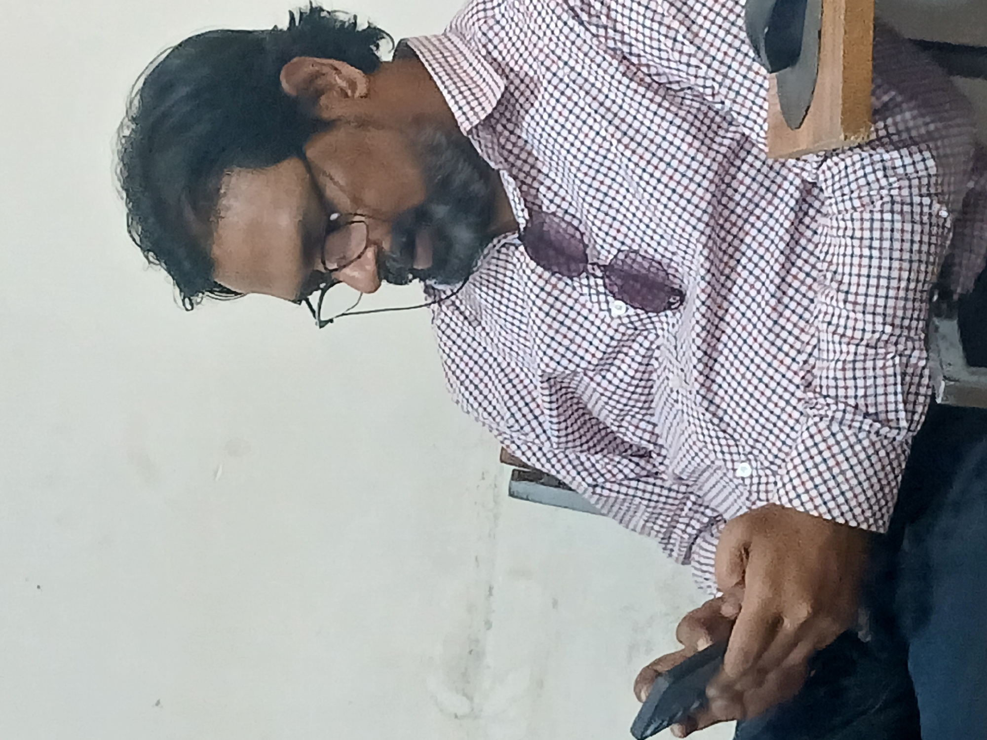 A man with dark hair and a beard is indoors. He is wearing glasses and a white and red checkered shirt. Sunglasses are hooked on his shirt. He is looking down and holding a dark colored cell phone. The location is Quetta City Tehsil, Pakistan.