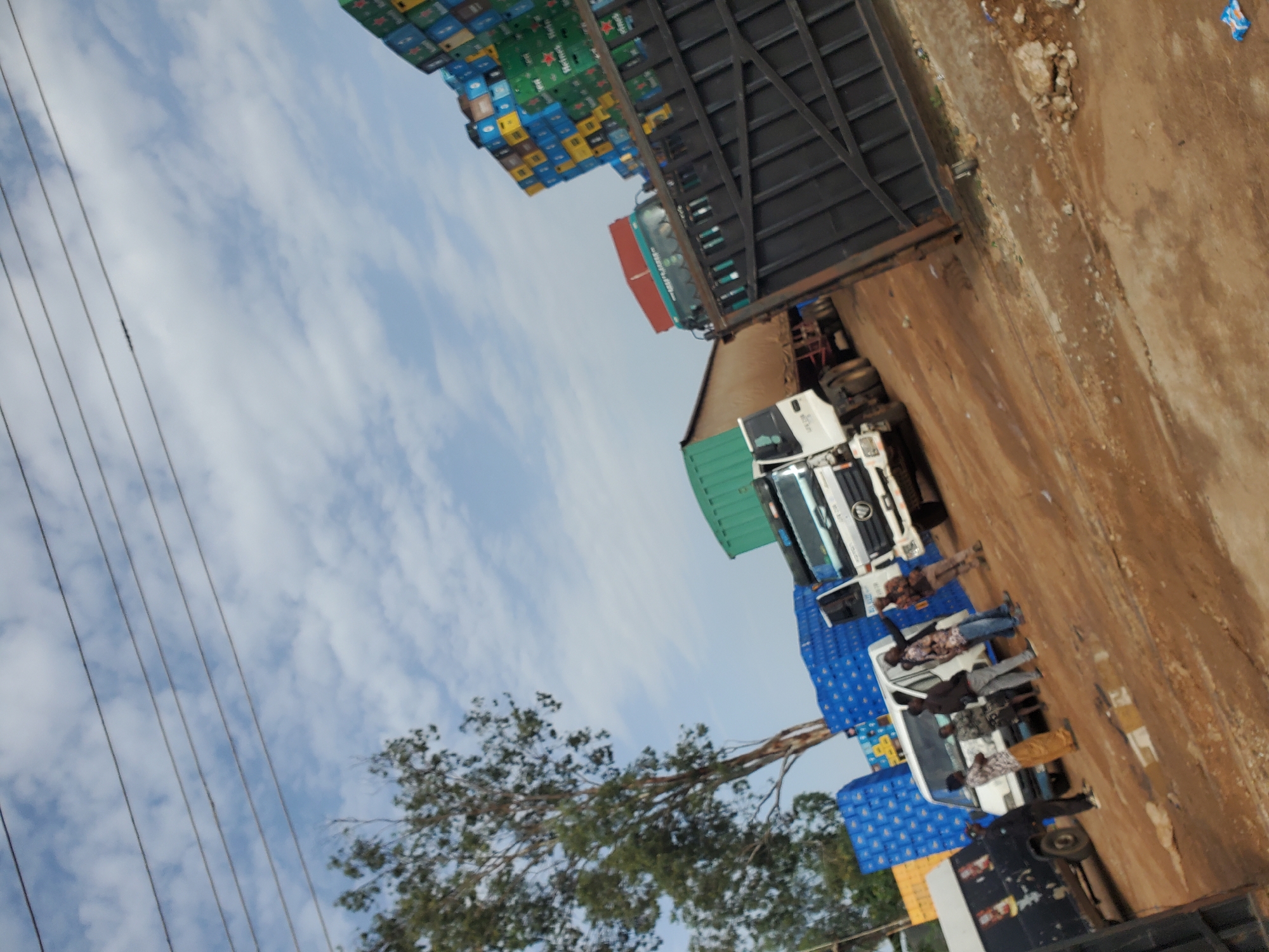 The image depicts an outdoor scene in Bukuru, Nigeria, possibly a distribution or loading yard. A fleet of vehicles is present, including a white delivery truck, a black van, and two larger trucks, one with a red trailer and another with a green cargo container. The vehicles are parked on a dirt ground with some puddles. Stacks of blue crates, possibly containing beverages, are visible behind some of the vehicles. In the foreground, a black metal gate stands open. Several individuals are interacting near the white delivery truck; one person is pointing, while others appear to be conversing. The sky is partly cloudy, and power lines are visible overhead. The overall impression is one of commercial activity, with goods being managed and transported.