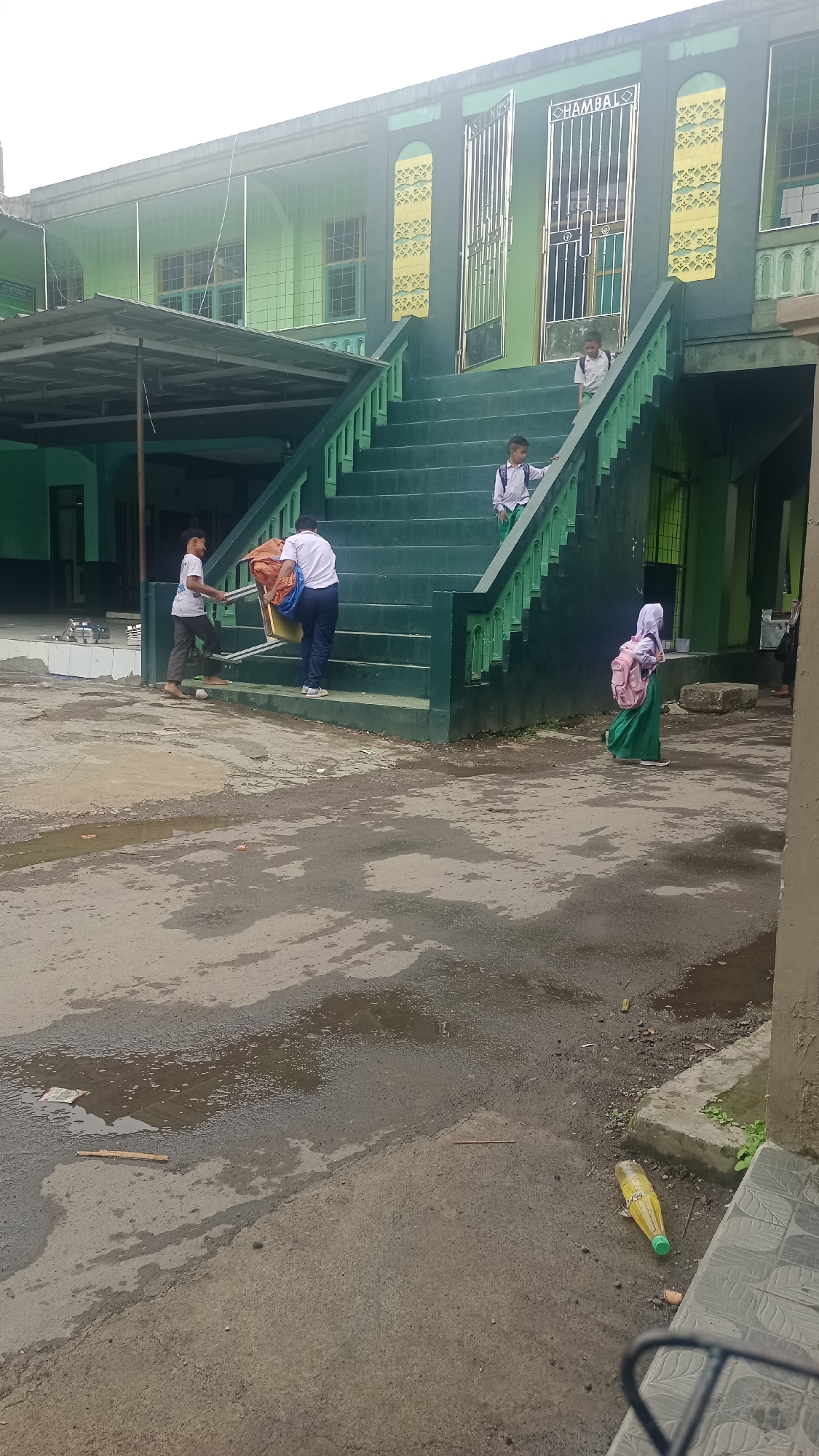 A group of children in school uniforms are near a green-painted building with a wide set of stairs. Two children are on the stairs, one in a white shirt and dark pants carrying large bags, and another in a white shirt and dark pants with a backpack. A third child, also in a white shirt and dark pants with a backpack, is on the stairs above. On the ground to the left, two boys in white shirts and dark pants are walking, one carrying a large bundle of orange and blue material. To the right, a girl in a white hijab and green dress with a pink backpack is walking away from the stairs. The building has decorative yellow elements and metal doors with the word "HAMBAL" visible. The ground in front of the building is wet with puddles and appears to be paved with asphalt. A yellow plastic bottle lies on the ground near a concrete step. The scene is outdoors, likely on school grounds in Karyamandala, Indonesia, given the architectural style and signage.