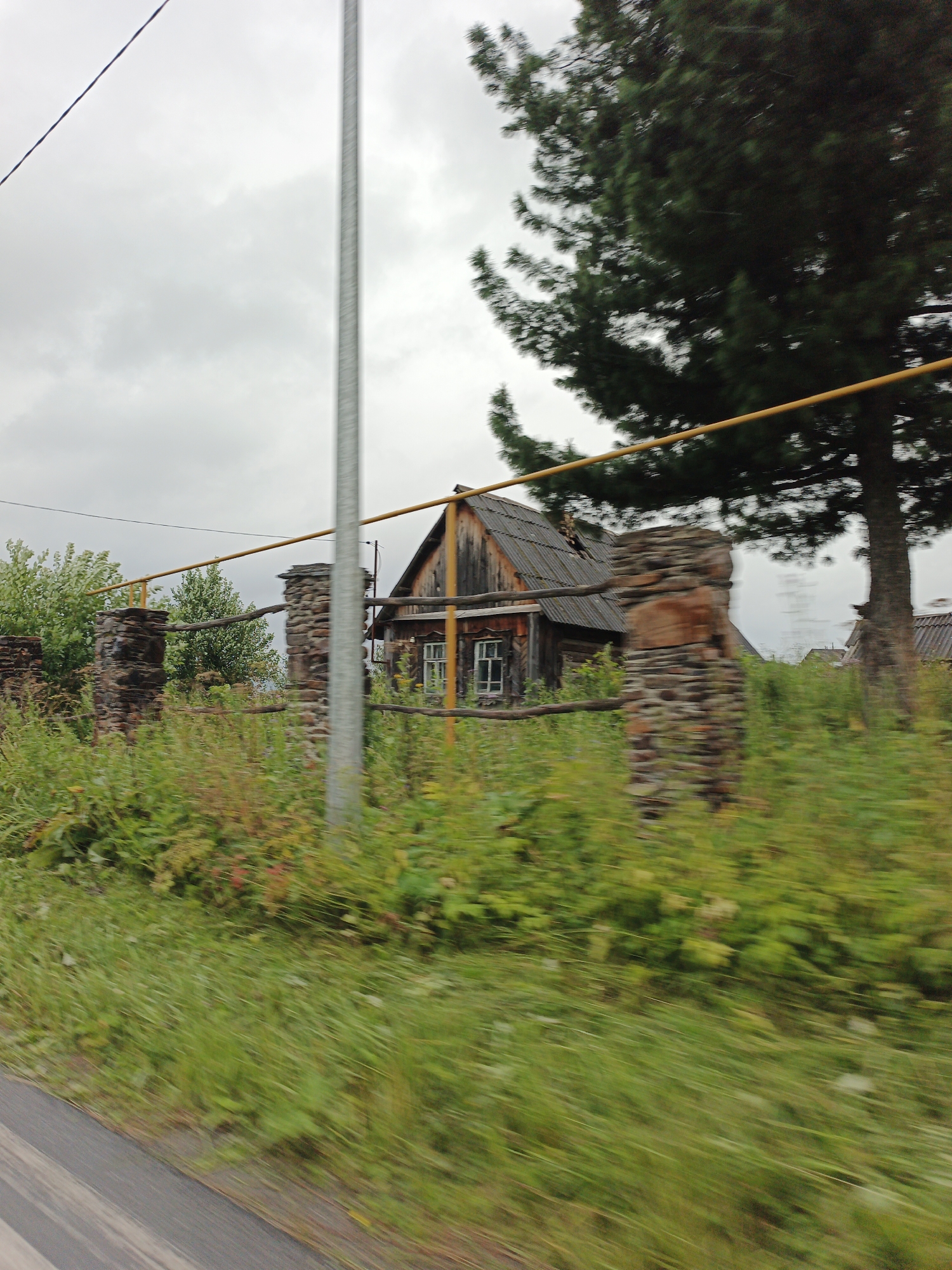 A photograph taken from a moving vehicle captures a rustic scene in Medvedyovka, Russia. In the foreground, blurred green grass and a grey, striped surface, likely a road, streak across the bottom of the frame. A tall, slender silver pole stands vertically on the right side of the image, bisecting the scene. Behind it, a wooden house with a dark, weathered exterior and a grey, corrugated roof is partially visible. A broken windowpane is evident on the left side of the house's facade. The house is bordered by a rustic fence constructed from rough-hewn logs and supported by two stone pillars. These pillars are made of irregularly stacked stones. A yellow pipe runs horizontally above the fence, supported by the stone pillars. A tall pine tree with dark green needles towers over the right side of the house, its trunk partially obscured. Lush green vegetation, with occasional hints of red, forms a dense border around the house and fence. The sky is overcast and a light grey, suggesting a cloudy or possibly rainy day. The overall impression is one of a rural, somewhat neglected property under a muted sky.