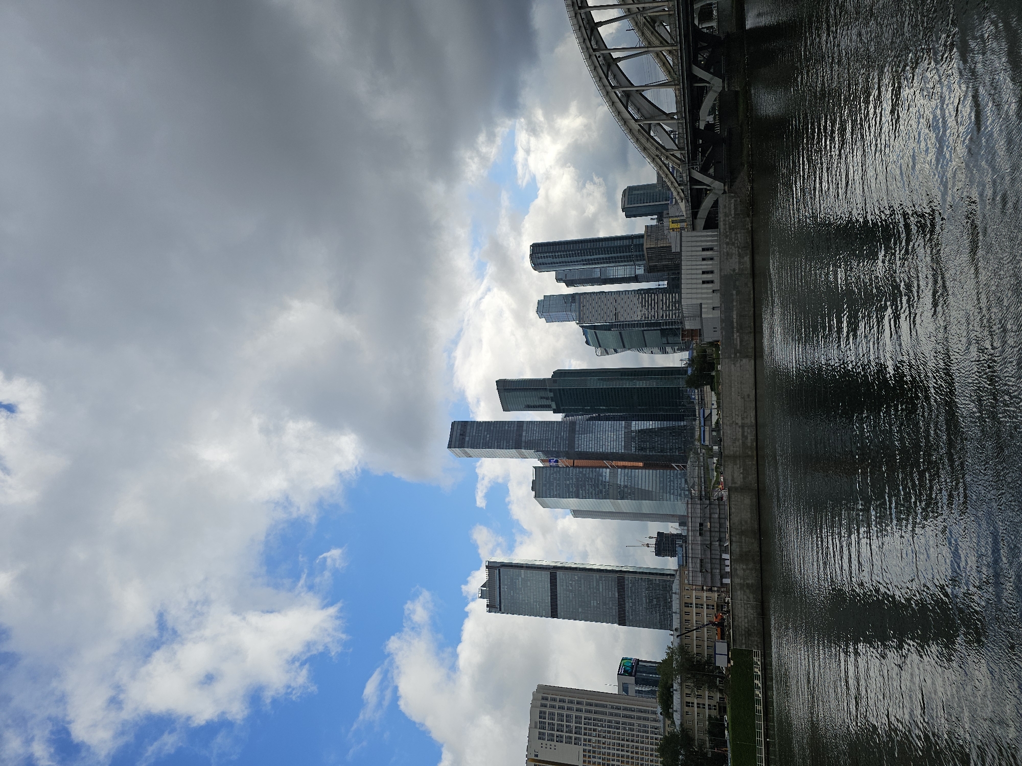 The image shows a view of the Moscow City business district with its modern skyscrapers, as seen from across the Moskva River. A large, arched bridge, likely a railway bridge, dominates the right side of the frame, with its complex steel structure and multiple arches visible. The skyscrapers in the background are a mix of glass and metal, reflecting the sky and surrounding environment. The sky is partly cloudy, with patches of blue sky visible amidst darker, more ominous clouds. The river in the foreground is dark and rippling, reflecting the buildings and the sky. The scene appears to be captured during the daytime, with overcast conditions contributing to a somewhat dramatic atmosphere. The image is a landscape orientation.