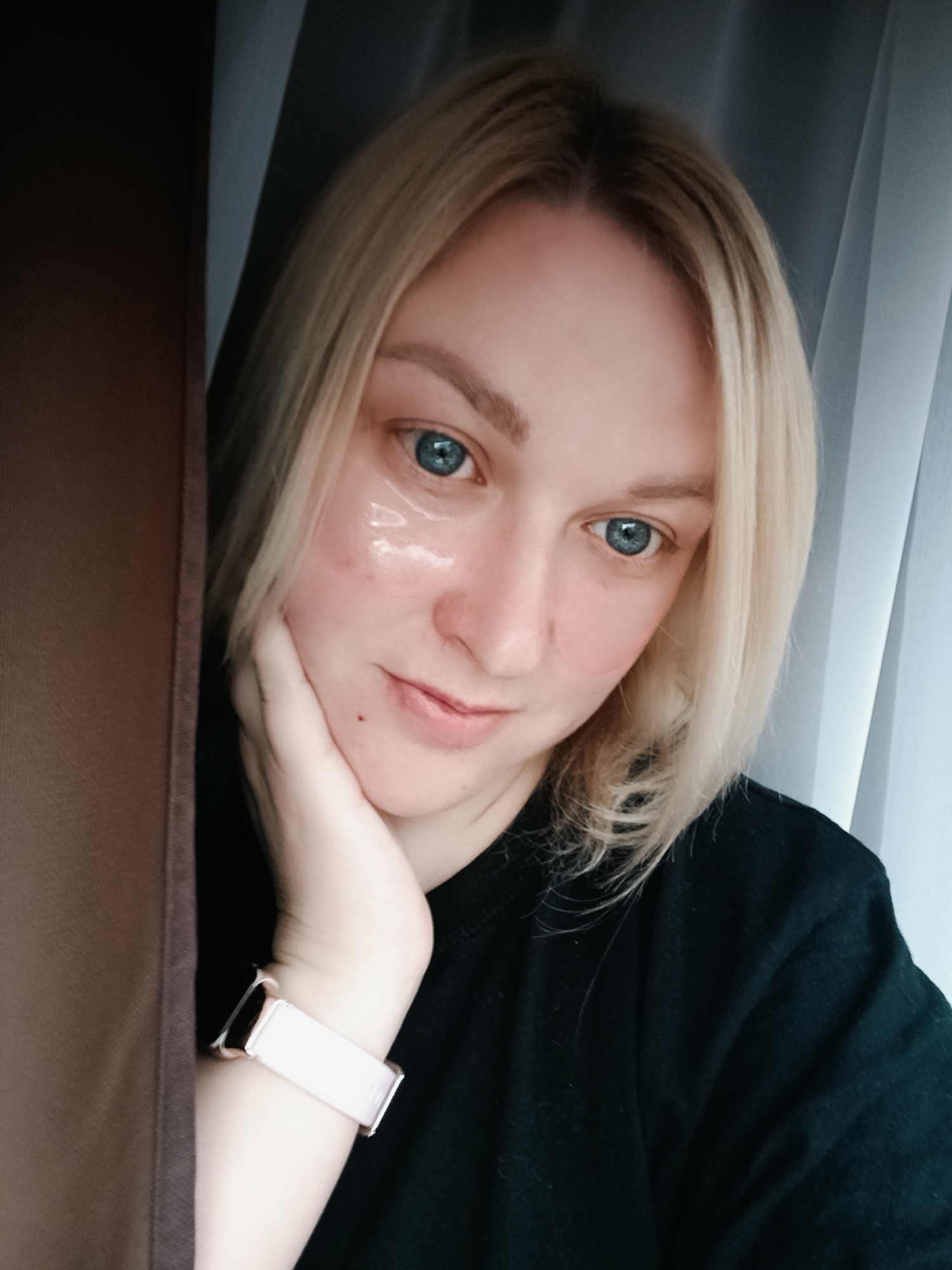 This is a medium close-up portrait of a young woman with blonde, shoulder-length hair and bright blue eyes, captured indoors in what appears to be Chaikovsky, Russia. She is looking gently towards the camera with a subtle, calm smile, resting her chin on her left hand. She wears clear, slightly pink-tinted hydrogel under-eye patches, suggesting a skincare or self-care routine. A light-colored (possibly rose gold or light pink) smartwatch is visible on her left wrist. She is dressed in a black t-shirt or top. The background features a dark brown curtain or fabric on the left side and a lighter grey/white curtain on the right, indicating an interior setting, likely near a window during daytime. The lighting is soft and natural. The woman's expression conveys a sense of calm and relaxation. No visible text is present in the image.