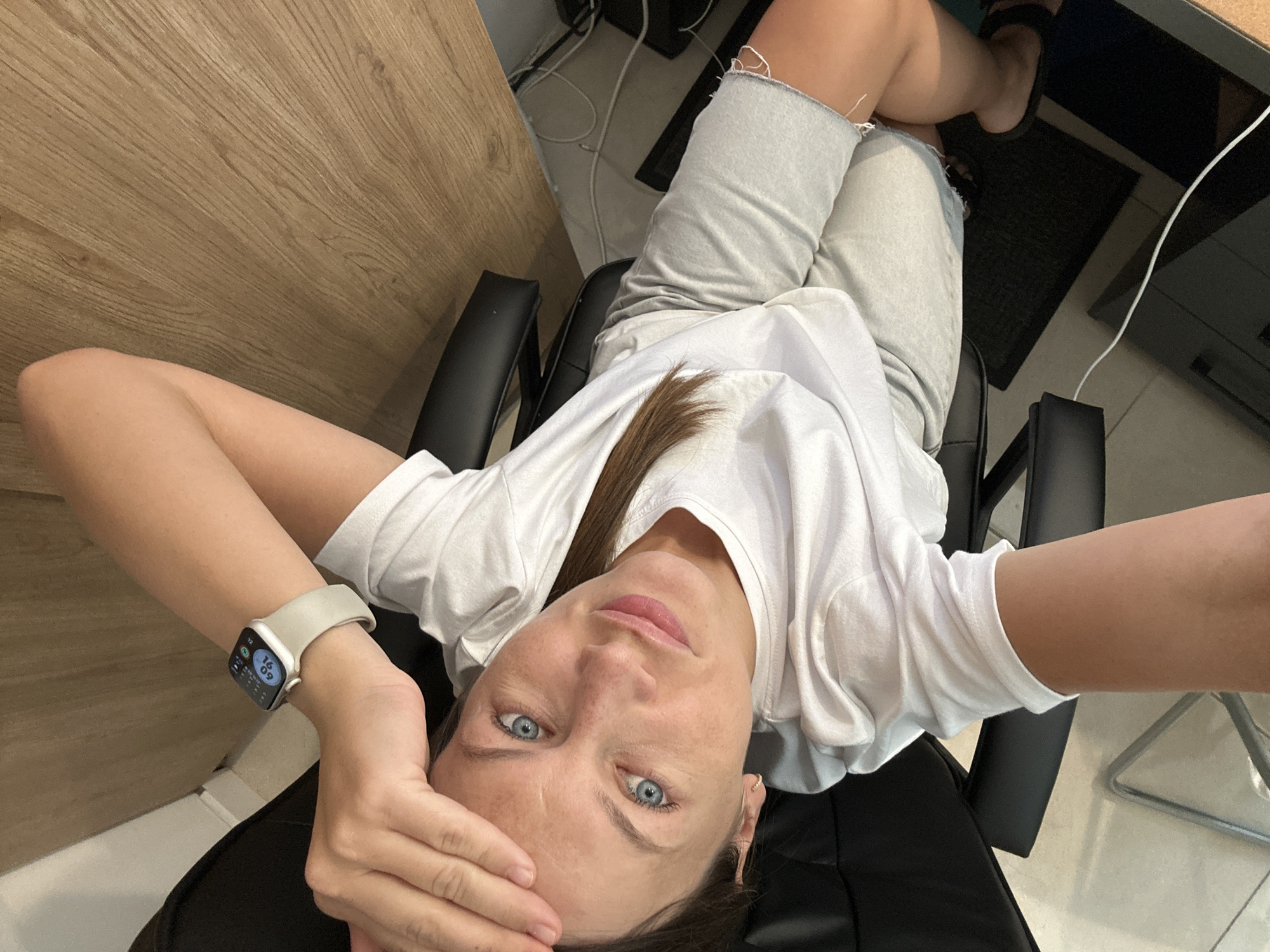 A woman with blue eyes and fair skin is lying on her side on a black office chair. She is wearing a white t-shirt and grey shorts. Her left arm is extended above her head, and her right hand is resting on her forehead. She is looking directly at the camera with a slight smile. A smartwatch is visible on her left wrist. The background shows a wooden desk, some electronic cables, and a black mat on the floor. The scene is indoors, possibly an office or home environment. There is no information in the image to suggest the location is in Boreti, Montenegro.