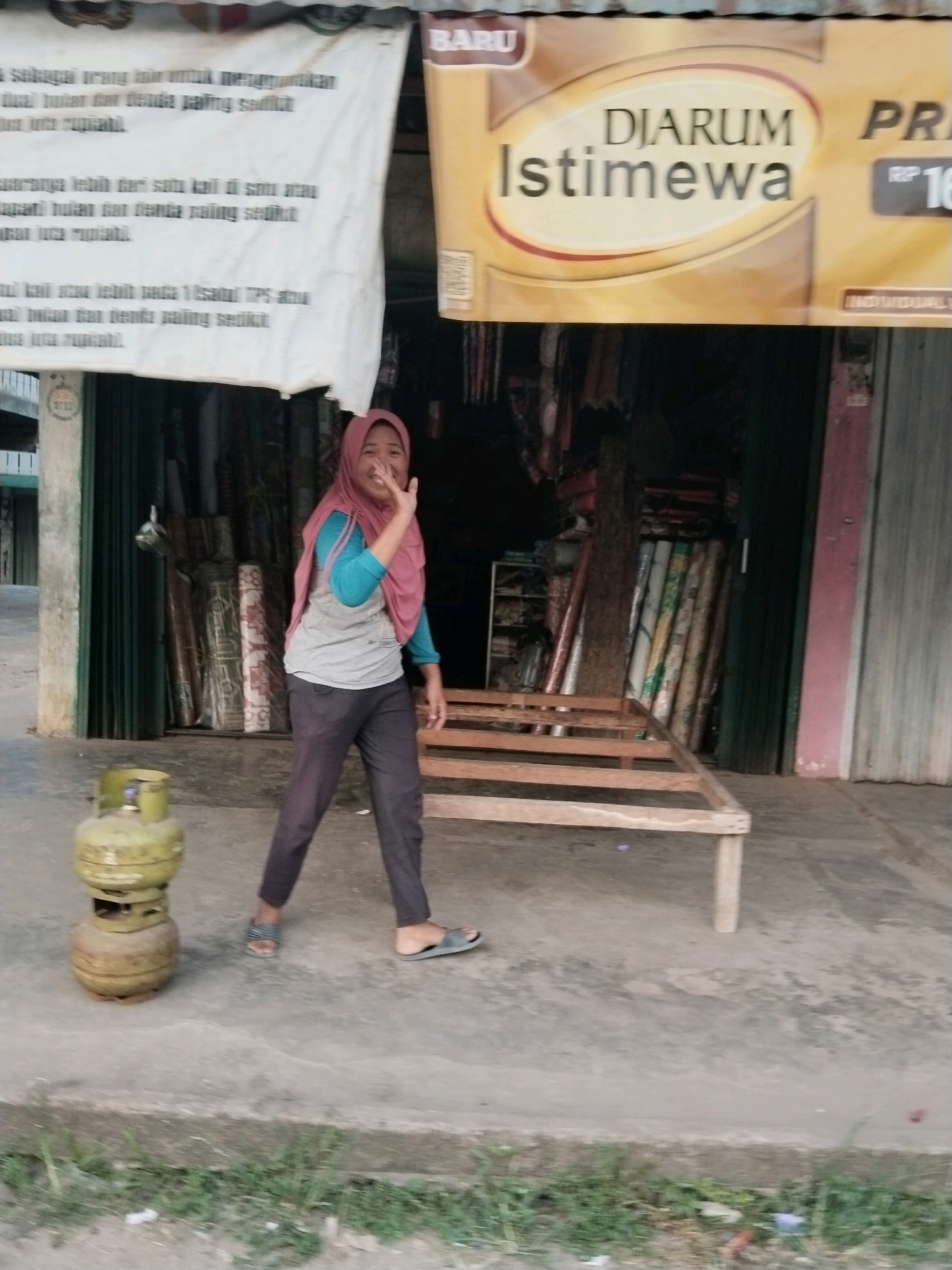 A young woman wearing a pink hijab and a gray t-shirt with a turquoise undershirt, dark pants, and sandals is walking on a paved surface. She is smiling and waving. To her left, there is a green propane tank. Behind her, there is a shop with green shutters and a wooden frame bed in front. A banner above the shop reads "DJARUM Istimewa". The environment appears to be a street in Suka Ramai, Indonesia, with buildings and some greenery visible.