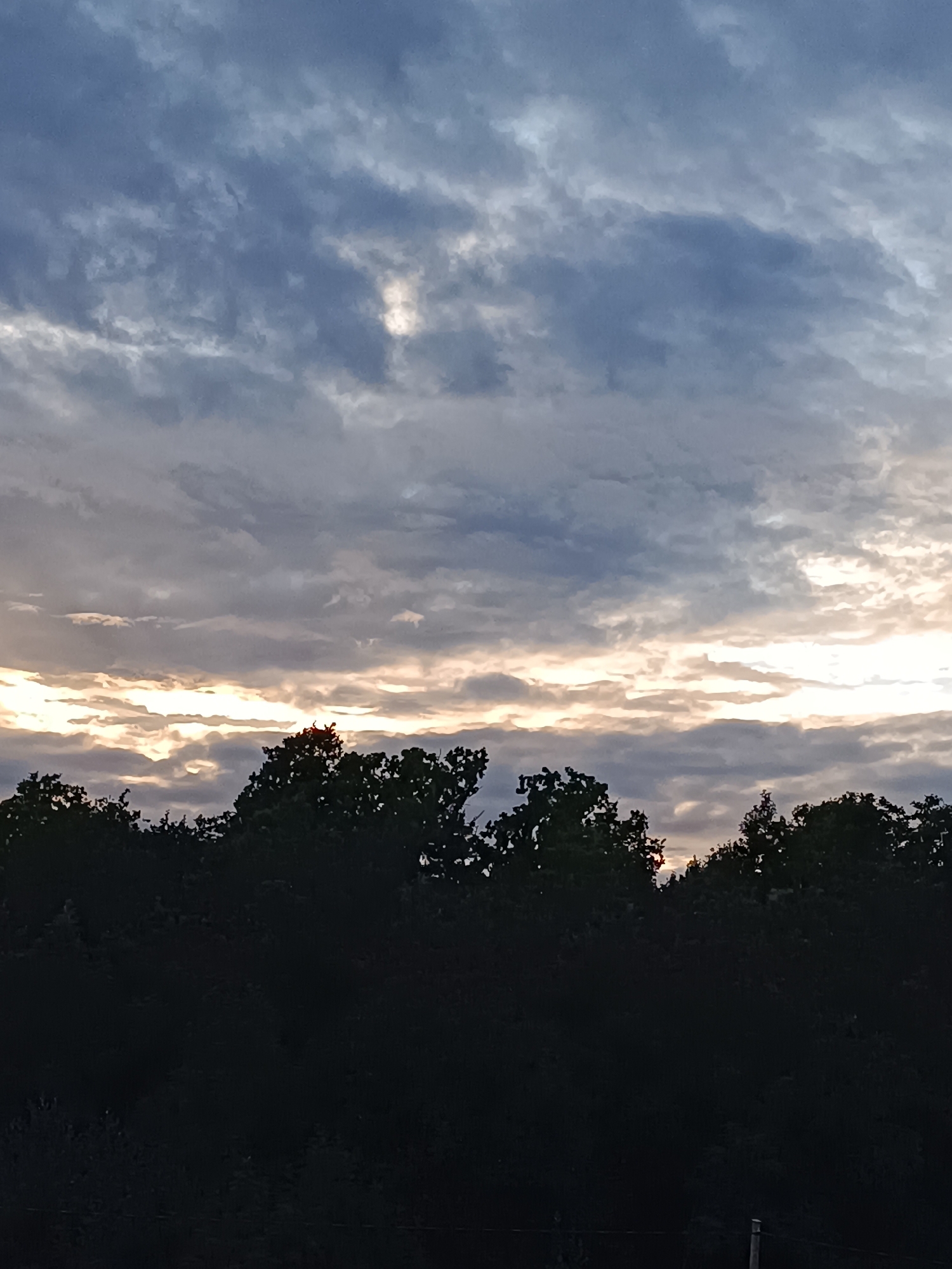 The image depicts a twilight sky over a treeline. The sky is filled with dramatic clouds, ranging from dark grey to lighter shades of blue and white, with streaks of bright, yellowish-orange light breaking through in the middle and upper portions, indicating the sun is low on the horizon. The treeline, silhouetted in dark green and black, spans the lower third of the image. The foliage is dense, with the tops of trees visible against the sky. No specific landmarks of Tebras, Latvia, are identifiable within the frame. The scene is devoid of any human presence or activity.