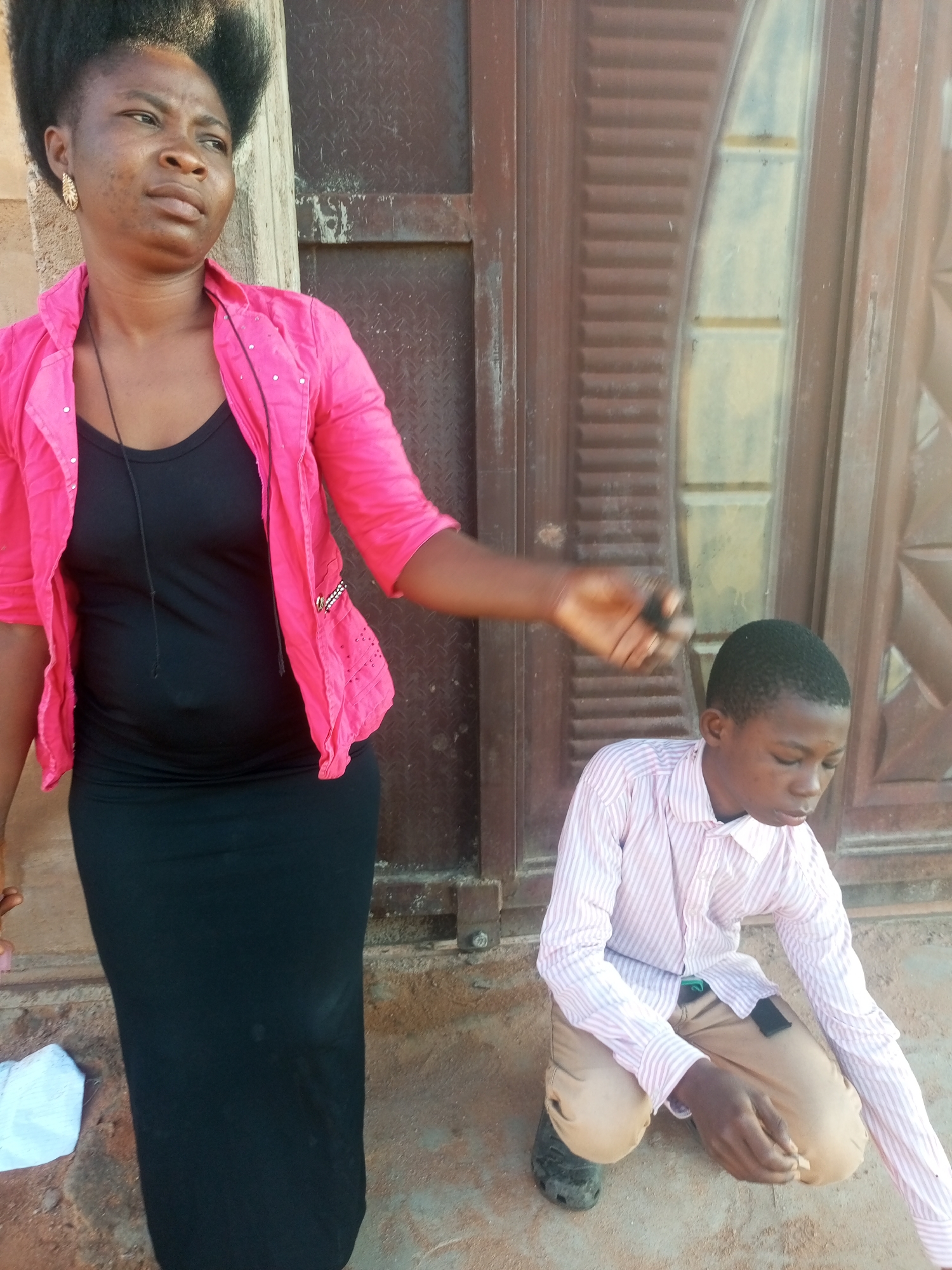 A woman in a pink jacket and black dress stands to the left of the frame, looking upwards and to her left. Her left hand is extended, holding a small black object. To her right, a young boy in a striped shirt and tan pants is kneeling on the ground, looking down. A brown wooden door is visible in the background. The scene appears to be outdoors, on a dirt ground, possibly in a compound in Tempure, Nigeria.
