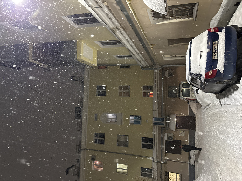 Snow falls heavily in a courtyard surrounded by buildings with illuminated windows. Two cars are parked close to a building, covered in snow. Air conditioning units are visible on the building’s exterior. A figure stands on the snowy ground, possibly looking up at the falling snow. The scene is captured at night with artificial lighting.