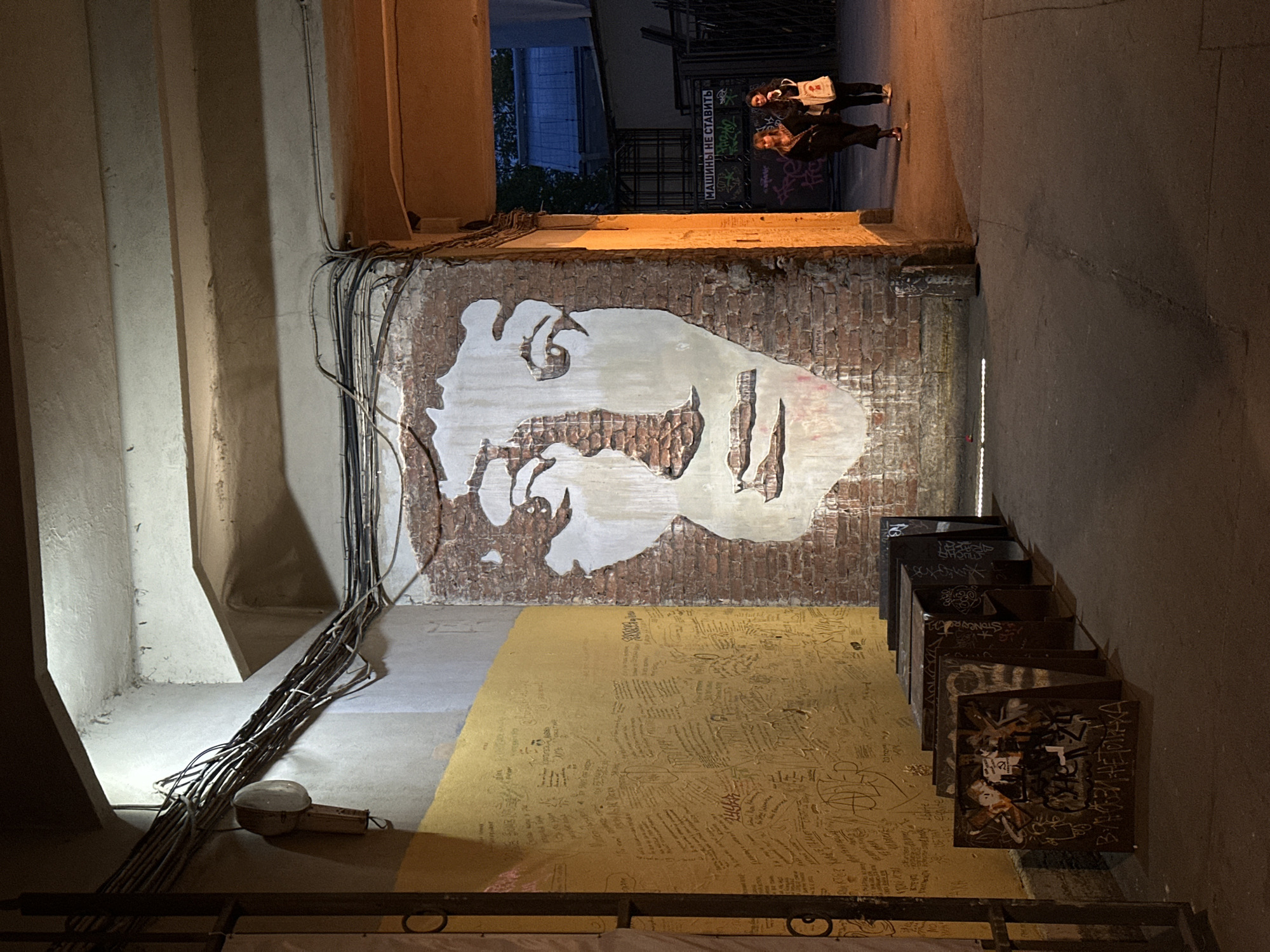 Captured from an elevated perspective, the photograph portrays the interior of an alleyway in an unknown city. The visual focus is a brick wall adorned with a stylized mural of a face, executed in white paint against the brick. The ground beneath the mural is coated in a yellow substance with dense, scribbled text.
A set of painted stairs leads downward on the right. The treads are covered in graffiti.
Electrical wiring is visibly routed along the left wall, leading down to the ground near the bottom left corner of the image. Above the mural, the alleyway opens up to the outside, where two individuals are discernible standing beneath a sign, facing away from the camera. The scene is dimly lit, emphasizing the mural.