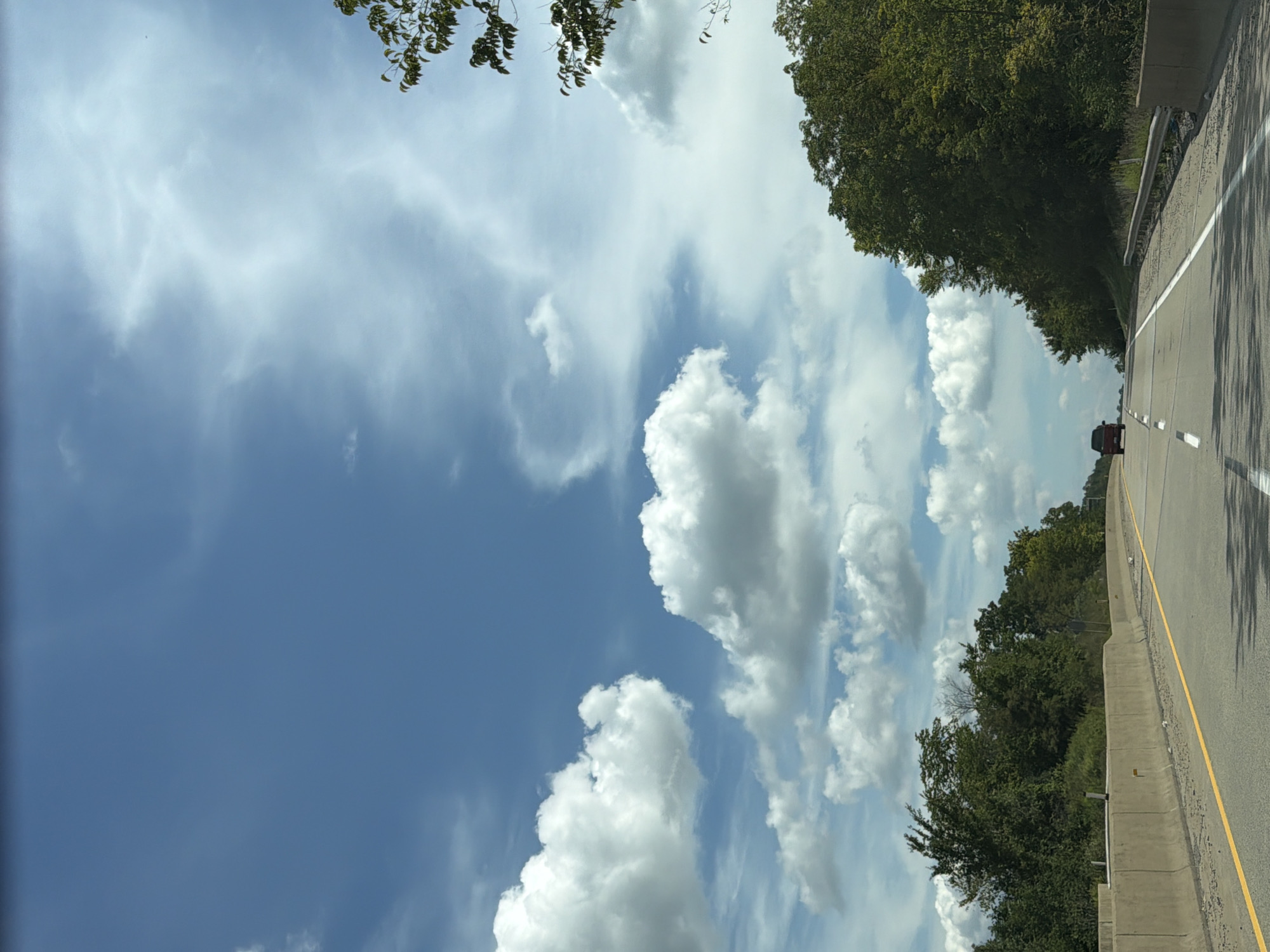 This is a landscape photograph taken from a vehicle traveling on a highway. The highway has a concrete barrier on the left and a guardrail on the right. The road is asphalt with yellow and white lane markings. The sky is a bright blue with scattered white cumulus clouds and some wispy cirrus clouds. Lush green trees line the sides of the highway. A dark red SUV is visible in the distance, traveling in the same direction. The scene appears to be set in a rural or suburban area, possibly North Fayette Township, United States of America, given the highway infrastructure and the presence of trees.