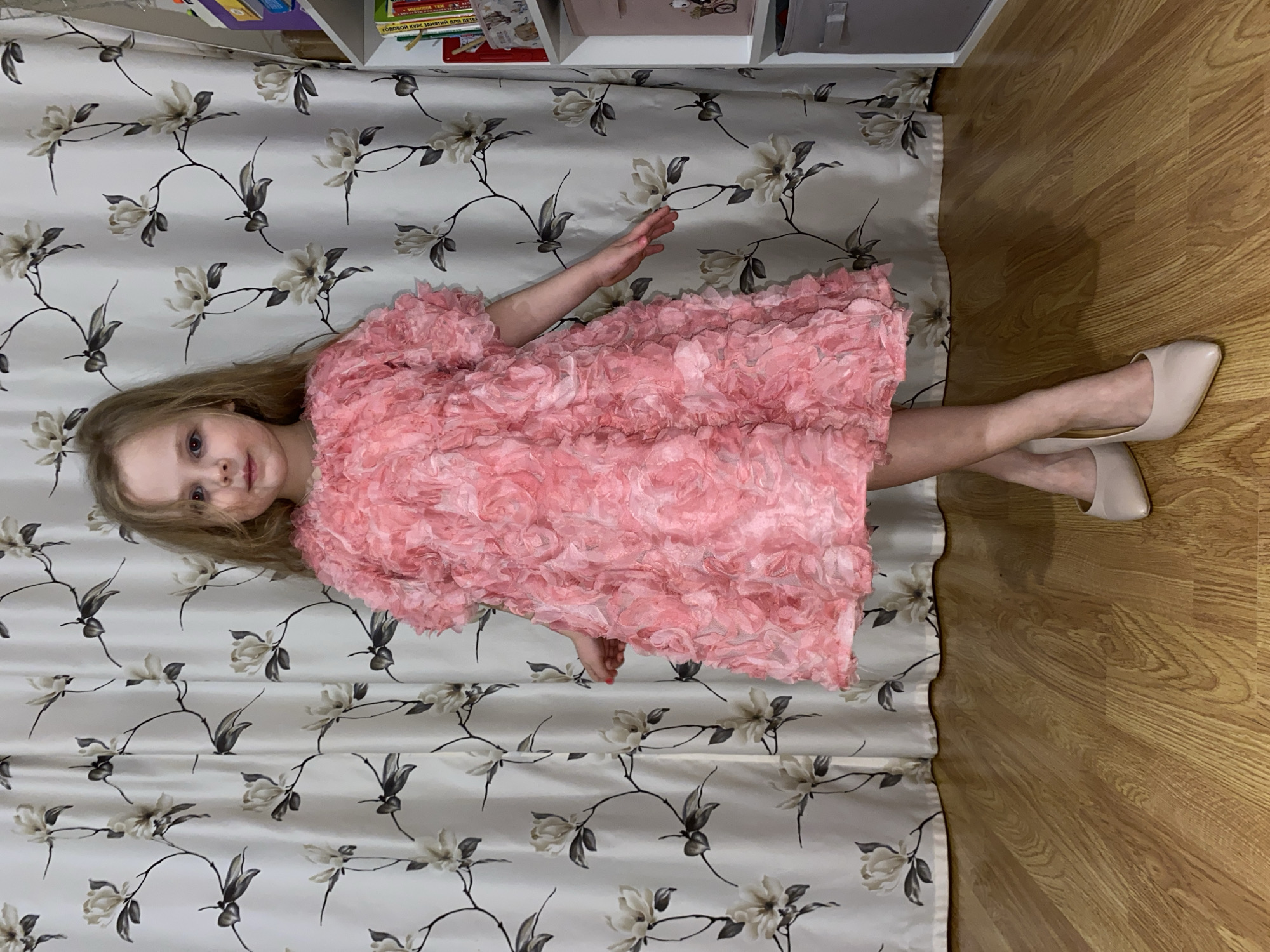 A young girl is posing indoors, standing in front of a patterned curtain and next to a wooden floor in Votkinsk, Russia. She has long, light brown or blonde hair and is wearing a voluminous, ruffled pink dress and light-colored high heels. The dress is entirely covered in layers of pink fabric ruffles or rosettes, giving it a textured, floral appearance.
Her feet are clad in beige or cream-colored pointed-toe high heels that appear several sizes too large for her, and her legs are crossed at the ankles. She stands with her left arm slightly extended outwards and looks directly at the camera with a neutral expression.
Behind her is a light-colored curtain with a repeating pattern of dark, winding stems and light, large blossoms, resembling magnolia flowers. To her right, a light-toned wooden floor is visible. Above the curtain, a partial view of a shelf unit or bookcase is seen, containing several books. Visible text on one of the books is in Cyrillic, reading "ГОДОВОЙ КУРС ЗАНЯТИЙ ДЛЯ ДЕТЕЙ" (Yearly Course of Classes for Children) and possibly "МИШКИНЫ СКАЗКИ" (Mishka's Tales). The scene depicts a domestic setting.