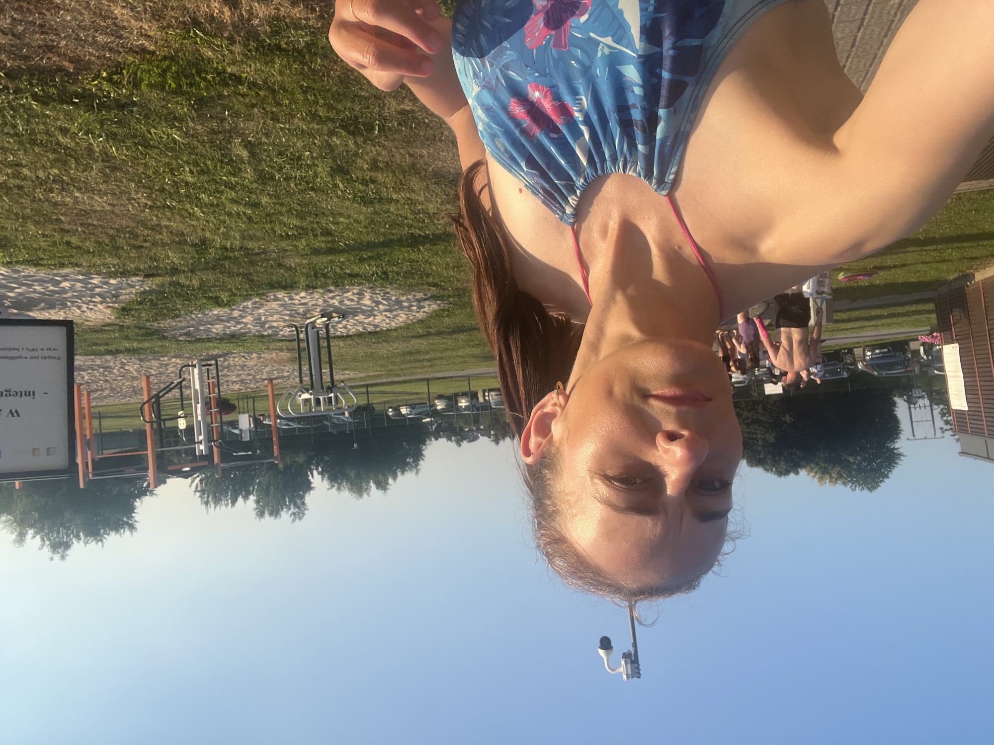 A woman, seen from the chest up, takes a selfie outdoors. She is wearing a blue and pink floral swimsuit and has long brown hair. She is looking at the camera with a neutral expression. Behind her, there is a grassy area with trees and an outdoor gym. In the background, there are cars parked and people walking. The sky is clear and blue. A security camera is visible mounted on a pole above and to the left of the woman's head.