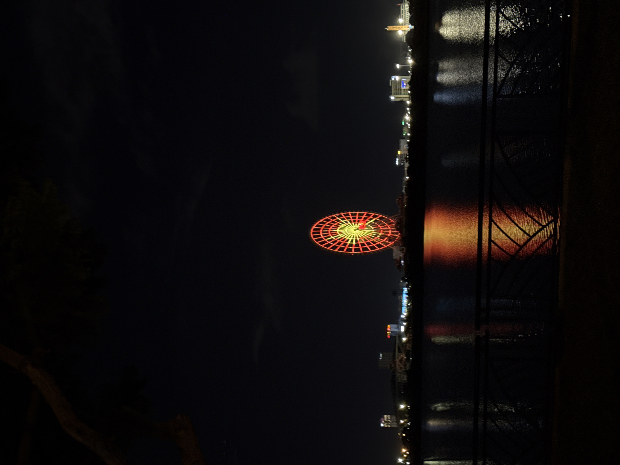 At night, a Ferris wheel in Đà Nẵng, Vietnam is illuminated with red and yellow lights. Across a body of water, a city skyline is visible with lights reflecting on the water. A window frame partially obscures the view on the right side of the image.