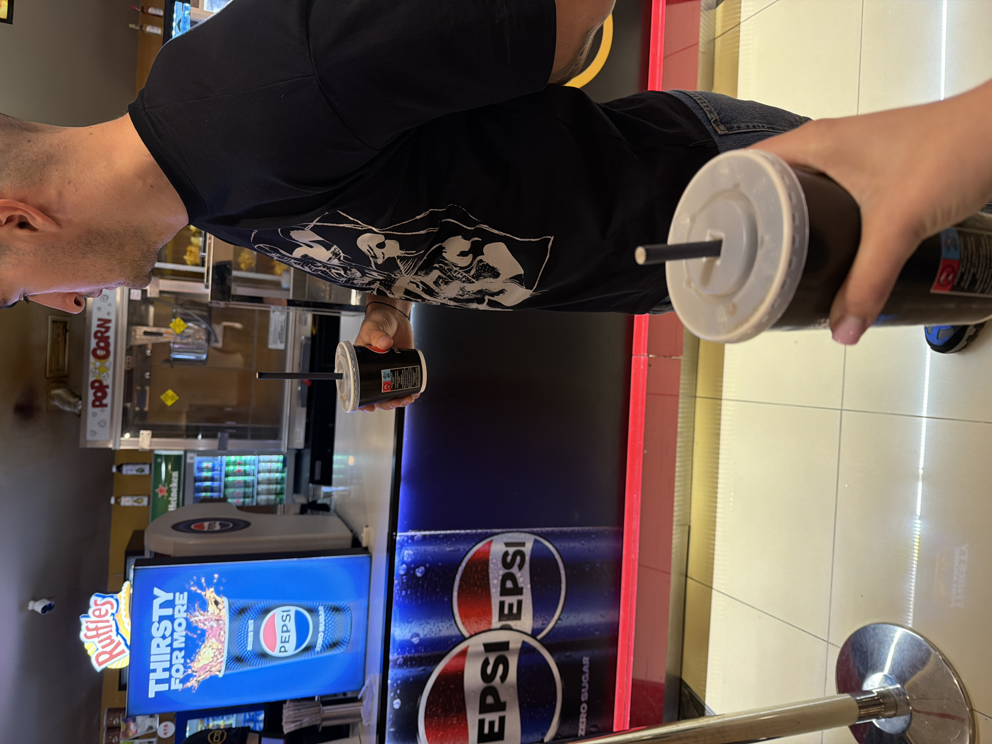 A medium, eye-level shot in a commercial setting. Two people stand at a counter, each holding a cup of Pepsi, with a Pepsi display behind the counter. The first person, a man, is wearing a black t-shirt and has closely cropped hair. A second person is visible in the frame holding a Pepsi drink.
Behind the counter are illuminated advertisements for Pepsi and Ruffles. A popcorn machine is also visible in the background. A drinks chiller is present behind the counter, full of Pepsi and Heineken beverages. The red and white circular logo of Pepsi is clearly displayed.
The location is a commercial establishment and a sign of "Popcorn" is visible in the background. The counter has a red trim.
Sofia, Bulgaria cannot be confirmed or denied with provided data.