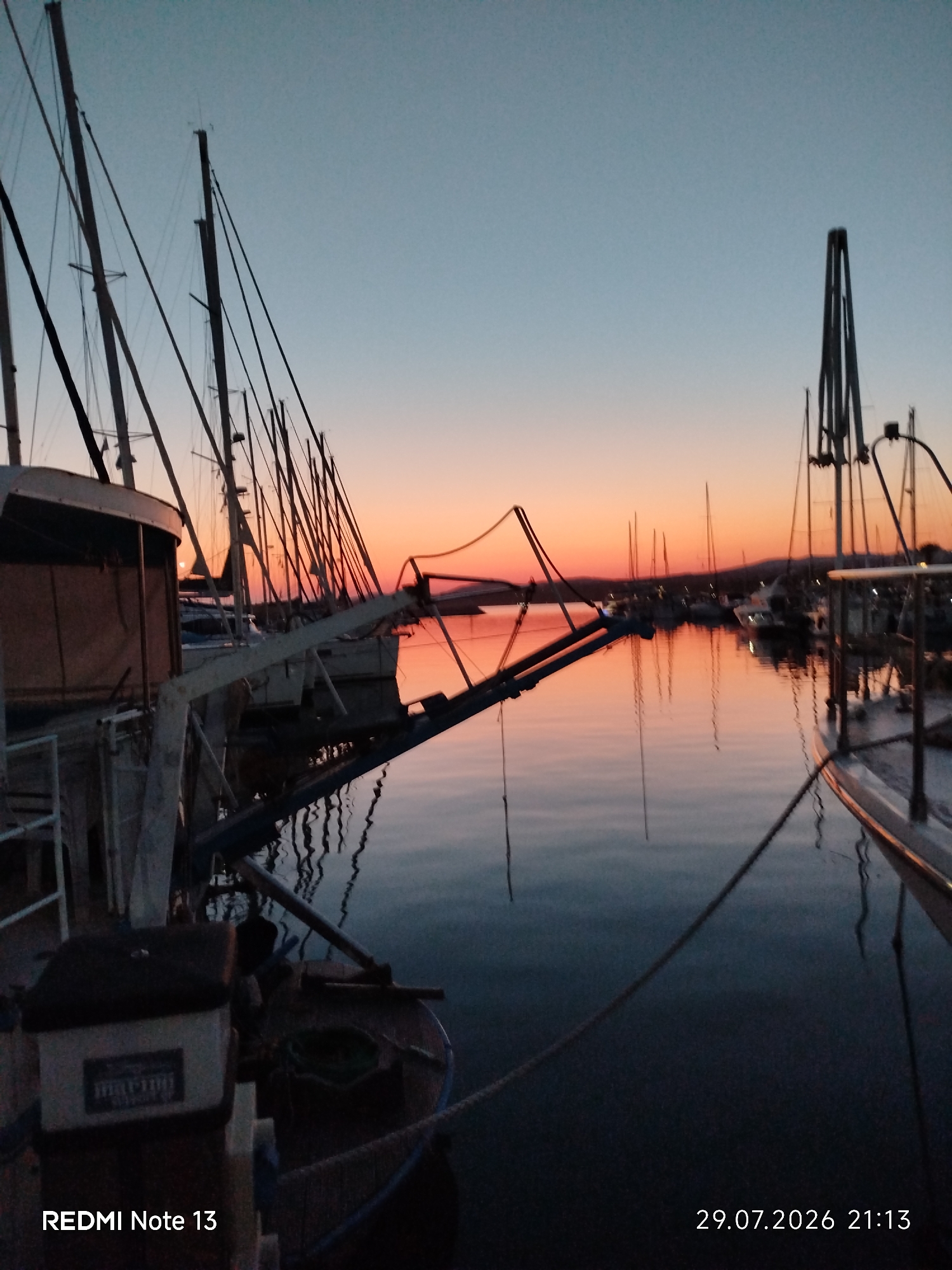 An outdoor, medium shot captures a harbor at dusk in Sithonia Municipal Unit, Greece. The sky displays a gradient from light blue to a warm orange near the horizon, indicative of a sunset. Silhouetted sailboats are docked, their masts creating a rhythmic pattern against the twilight sky. The calm water reflects the sky's colors and the boats, creating a mirror image. In the foreground, the dark hull of a boat occupies the lower right portion of the frame, with a rope extending across the water. A white storage container with a label is visible on the left. The bottom left corner of the image displays "REDMI Note 13" and the bottom right corner shows "29.07.2026 21:13". The overall atmosphere is serene.