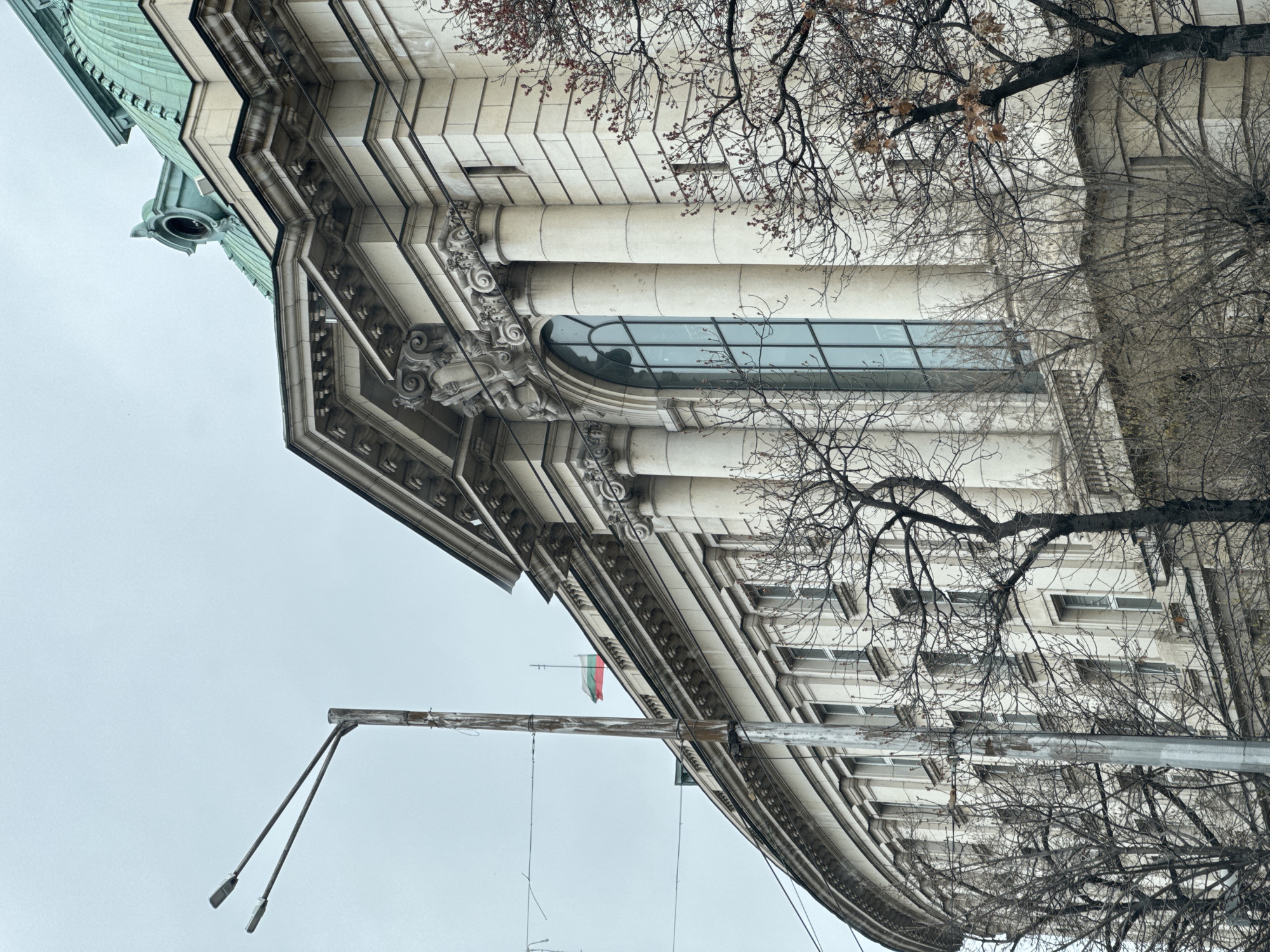 The image depicts a building in Sofia, Bulgaria, viewed from a low angle, looking up. The building's facade is white with classical architectural details, including columns, moldings, and a large arched window. The roof appears to be green. Bare trees are visible in the foreground. A metal street lamp pole extends diagonally across the lower part of the frame, and the Bulgarian flag hangs from the building. The sky is overcast and gray. The location is likely a government building in central Sofia due to its architectural style and the presence of the Bulgarian flag.