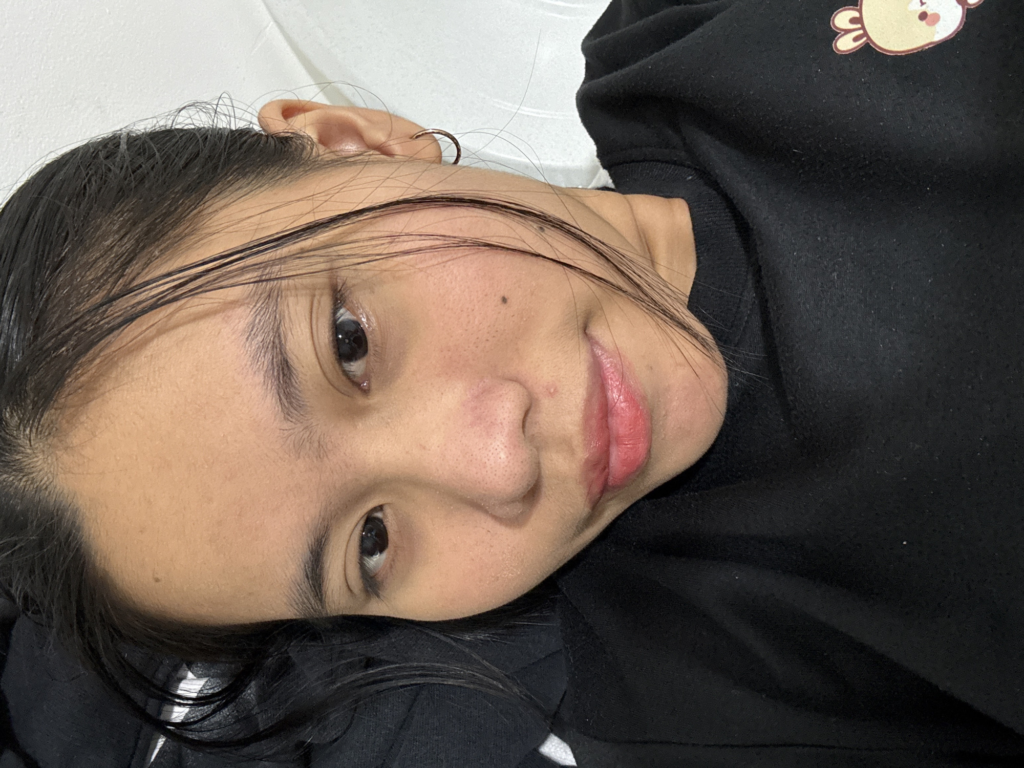 A close-up, selfie-style photograph shows a young woman with dark hair, pulled back but with several strands falling forward. She is wearing a black crew-neck sweatshirt with a cartoonish illustration of a light-brown bear on the right side of the chest. The woman is lying down, with the camera angled slightly from above. Her head is tilted to her left, and she is looking directly at the camera with a neutral expression. Her skin appears to have visible pores, especially on her nose and cheeks, and there is a small mole on her right cheekbone. Her lips are painted a light pinkish-red. A thin, gold hoop earring is visible in her right ear. The background consists of a white, textured surface and a darker, soft surface on the left side of the frame, possibly a pillow or blanket. The lighting is bright and diffused. The image was taken indoors.