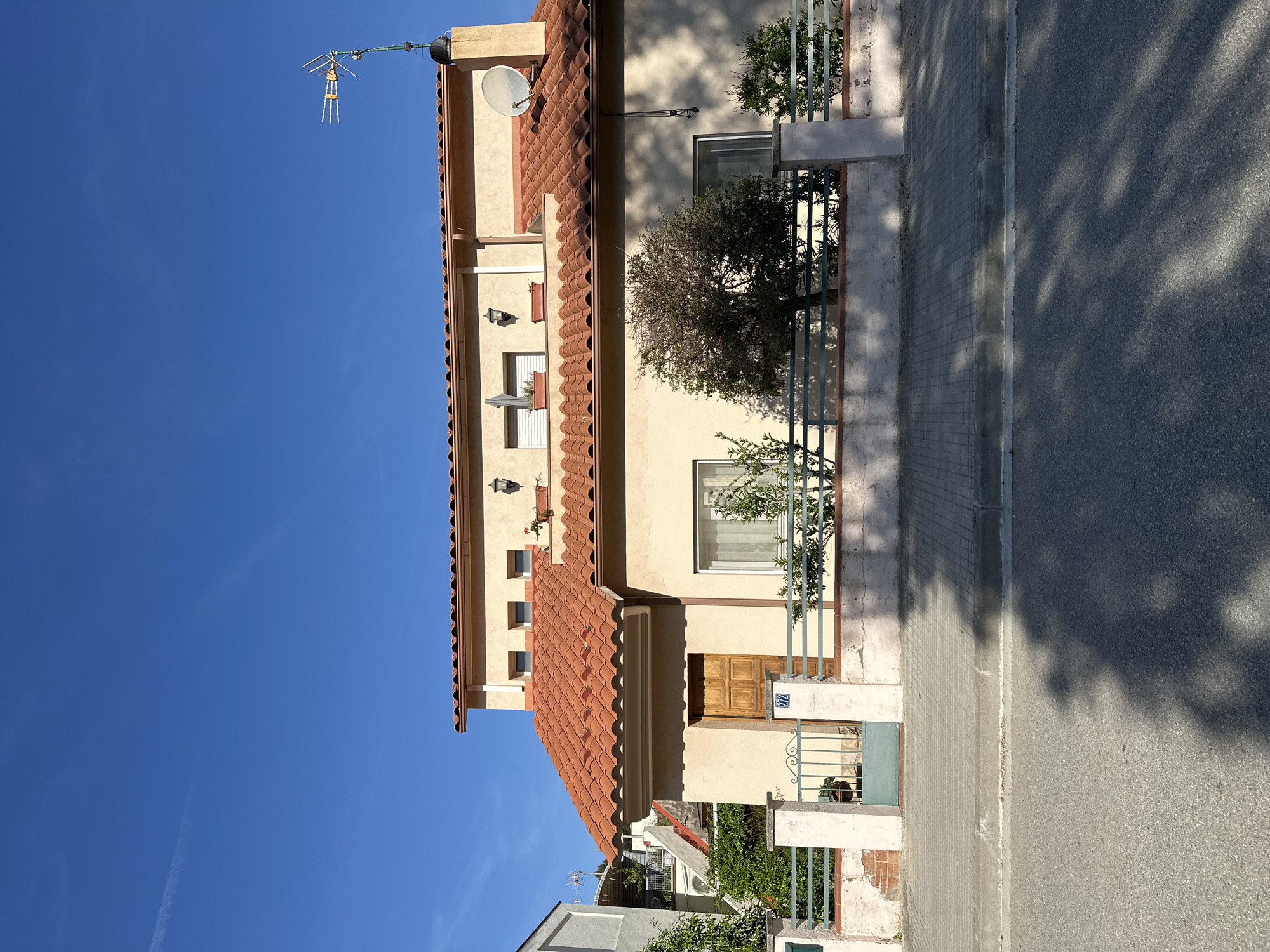 The image, rotated 90 degrees clockwise, captures a sunny residential street scene. A two-story house with a light-colored facade and a distinctive terracotta tile roof dominates the view. On the roof, both a traditional television antenna and a white satellite dish are present.
The house features multiple windows; some are fitted with closed white shutters, and one on an upper floor holds a planter with small red flowers. A wooden front door is visible near the entrance. In front of the property, a green metal fence and gate are partially visible, surrounded by lush green trees and bushes.
The clear, bright blue sky and the long, dappled shadows cast by trees on the asphalt road and paved sidewalk indicate a sunny day, likely late morning or early afternoon. The architectural style, particularly the terracotta roof, is characteristic of buildings found in Mediterranean regions, consistent with the location of La Garriga, Spain.
Visible text includes the number "77" on a blue house number plaque attached to the wall, and the number "177" on a white plate affixed to the green metal gate.