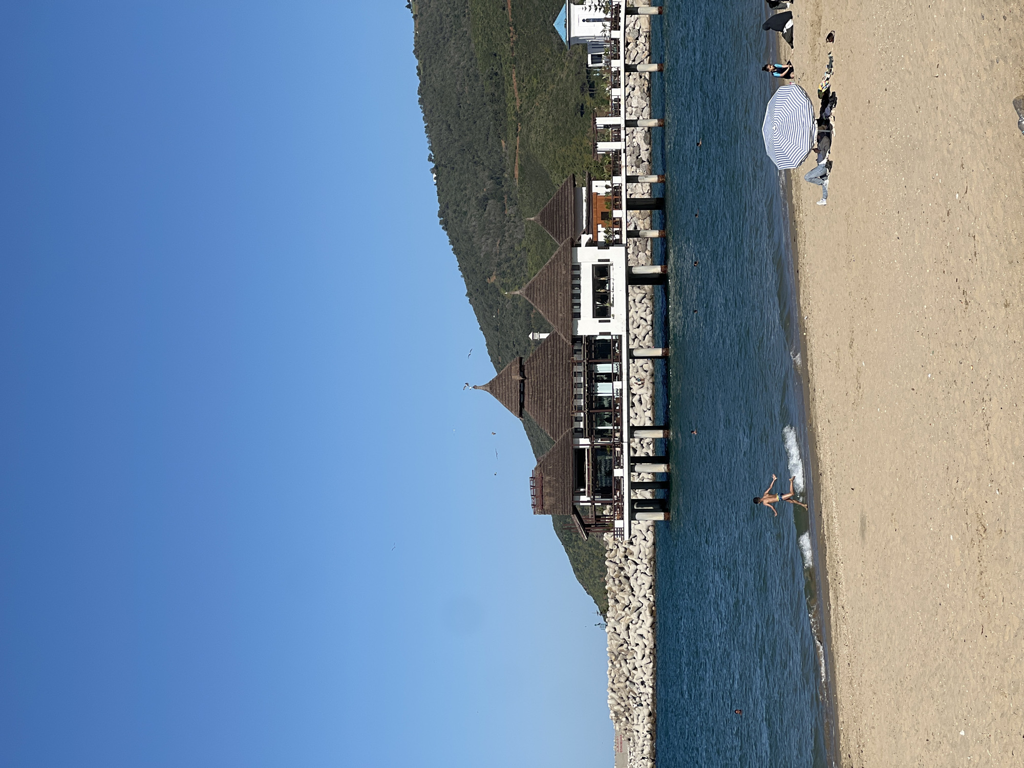 The image depicts a beach scene in M'Diq, Morocco. The beach is sandy, with clear blue water meeting the shore. A person is floating in the water near the shoreline. Further down the beach, there is a group of people under a blue and white striped umbrella. To the left, a structure on stilts extends into the water. The structure appears to be a multi-story building with a dark roof and white walls. A densely vegetated hill rises behind the building. The sky is clear and blue. Concrete barriers form a breakwater along the coastline.