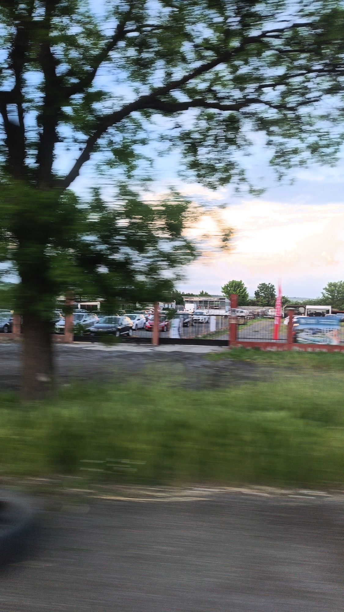 The image depicts a scene from a moving vehicle. A large tree with green leaves is in the foreground. Behind the tree, there is a parking lot filled with numerous cars. A building with a sign that reads "TOKOHMAPMHTHOH" is visible in the background. A red fence surrounds the parking lot, and advertisements or promotional materials are attached to it. The sky is overcast with a mix of blue and white. The scene appears to be captured on a roadside in Burgas, Bulgaria. The photo is blurry due to the movement of the vehicle.