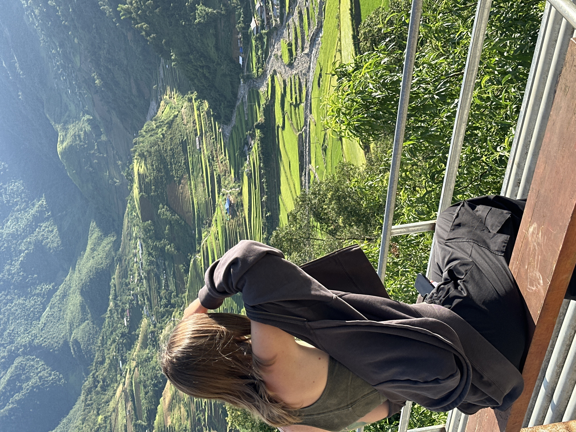 A person is observed from behind, seated on a light brown wooden bench, gazing over a vast mountainous valley in Tú Lệ, Vietnam. The individual has medium-length brown hair and is dressed in an olive green sleeveless top, with a dark grey hooded jacket draped over their shoulders, and dark grey cargo pants. Their right hand is raised near their forehead, and a black smartphone rests on their lap. A grey metal railing and lush green foliage occupy the immediate foreground. The valley below features extensive bright green rice terraces carved into the slopes of verdant mountains. A wide, stony riverbed with visible water channels winds through the valley floor. Scattered small buildings and houses, some with blue roofs, are visible among the terraces and alongside the river. Strong sunlight illuminates the scene, creating bright areas and distinct shadows across the landscape.