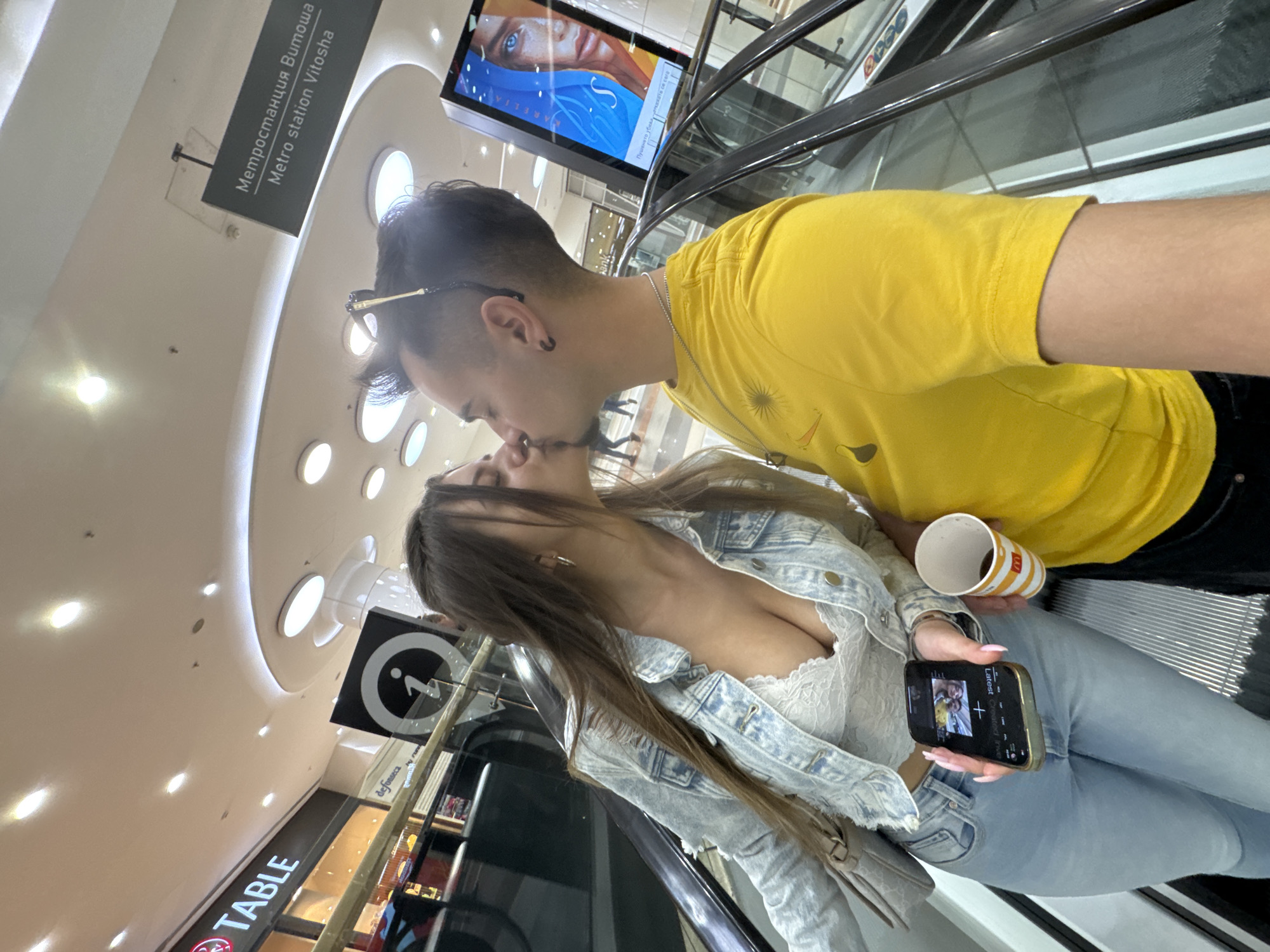 Here's a detailed description of the image:
**Scene:** The image captures a close-up shot of a young couple kissing on an escalator inside a shopping mall in Sofia, Bulgaria. The presence of signs in Bulgarian, specifically mentioning "Метростанция Витоша" and "Metro station Vitosha", indicates the location. The background reveals elements of a modern shopping mall interior, including ceiling lights, store signs (e.g., TABLE), and a digital advertising screen.
**People:**
* A young man with dark hair, wearing a yellow t-shirt and sunglasses.
* A young woman with long brown hair, wearing a white lace top and a light blue denim jacket, holding a mobile phone and a paper cup.
**Objects:**
* Escalator
* Mobile phone displaying an image
* Paper cup (likely from McDonald's)
* Mall signage (e.g., "Метростанция Витоша", "Metro station Vitosha", TABLE, information symbol)
* Digital advertising screen
* Storefronts
* Ceiling lights
**Actions and Interactions:**
* The couple is kissing.
* The woman is holding a mobile phone and a paper cup.
**Environment:**
* The setting is inside a modern shopping mall, likely near an entrance or exit leading to the Vitosha Metro station in Sofia. The presence of advertising screens, shops, and escalators confirms this.