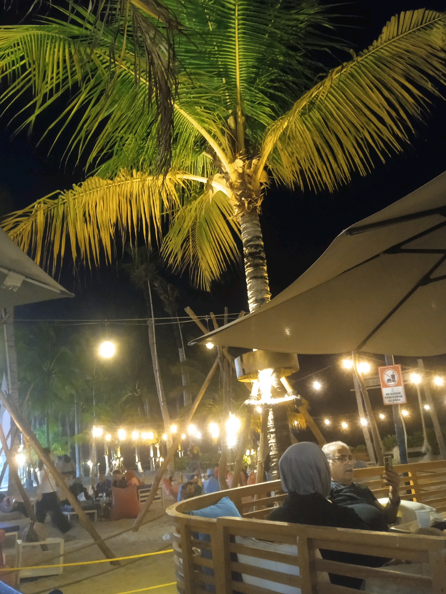 The image depicts an outdoor dining area at night, likely a beachside cafe or restaurant, in Kosambi Barat, Indonesia. A large palm tree with green and yellow fronds dominates the foreground, illuminated from below, casting shadows. Several tables and seating areas are visible, some with people seated. In the mid-ground, strings of warm-toned lights are strung between poles, creating a festive ambiance. Two individuals are seated in the foreground on the right, facing away from the camera; one is wearing a gray hijab and the other a black t-shirt and glasses, looking at a smartphone. A sign with a crossed-out food and drink symbol is mounted on a pole behind them. In the background, more palm trees and lights are visible, suggesting a tropical location. The overall atmosphere is relaxed and communal, with a focus on outdoor leisure.