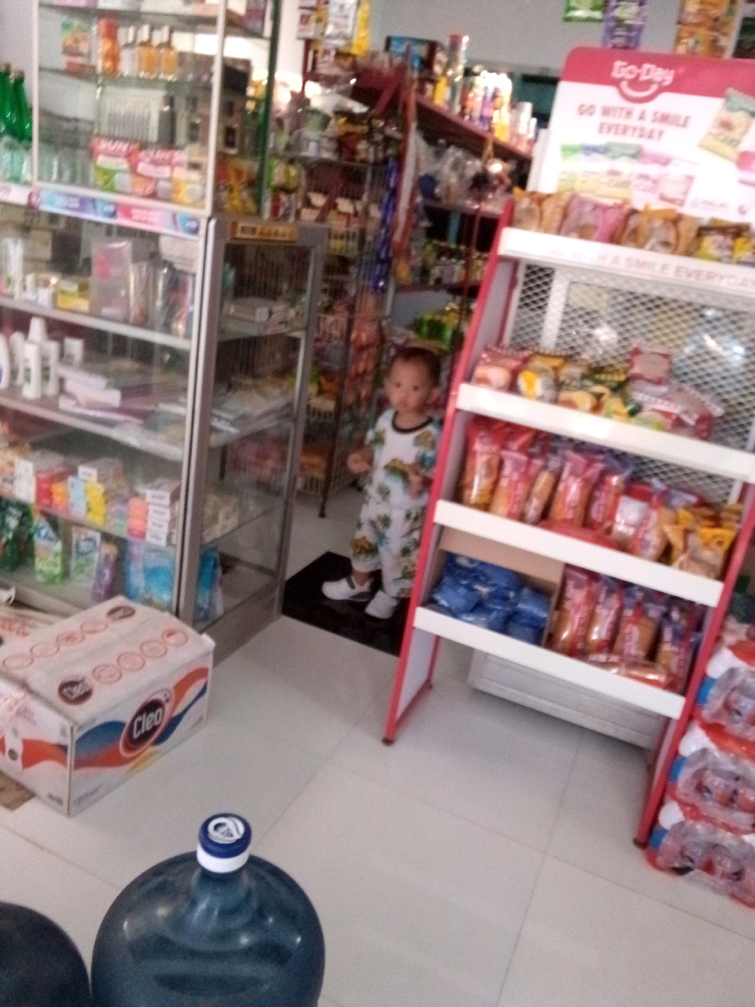 The image depicts a small convenience store or minimarket located in Simo, Indonesia. A young child, wearing a white outfit with a playful pattern, stands in the aisle between shelves stocked with various products. To the left, glass-fronted shelves display an assortment of packaged goods, including what appear to be cleaning supplies, toiletries, and food items. A prominent cardboard box labeled "Cleo" is visible in the foreground on the left. To the right, a display rack is filled with packaged snacks and beverages. In the background, a larger display board with the "Go-Day" logo and text is mounted. The floor is tiled, and two large blue water cooler bottles are partially visible in the lower portion of the image. The lighting suggests an indoor environment.