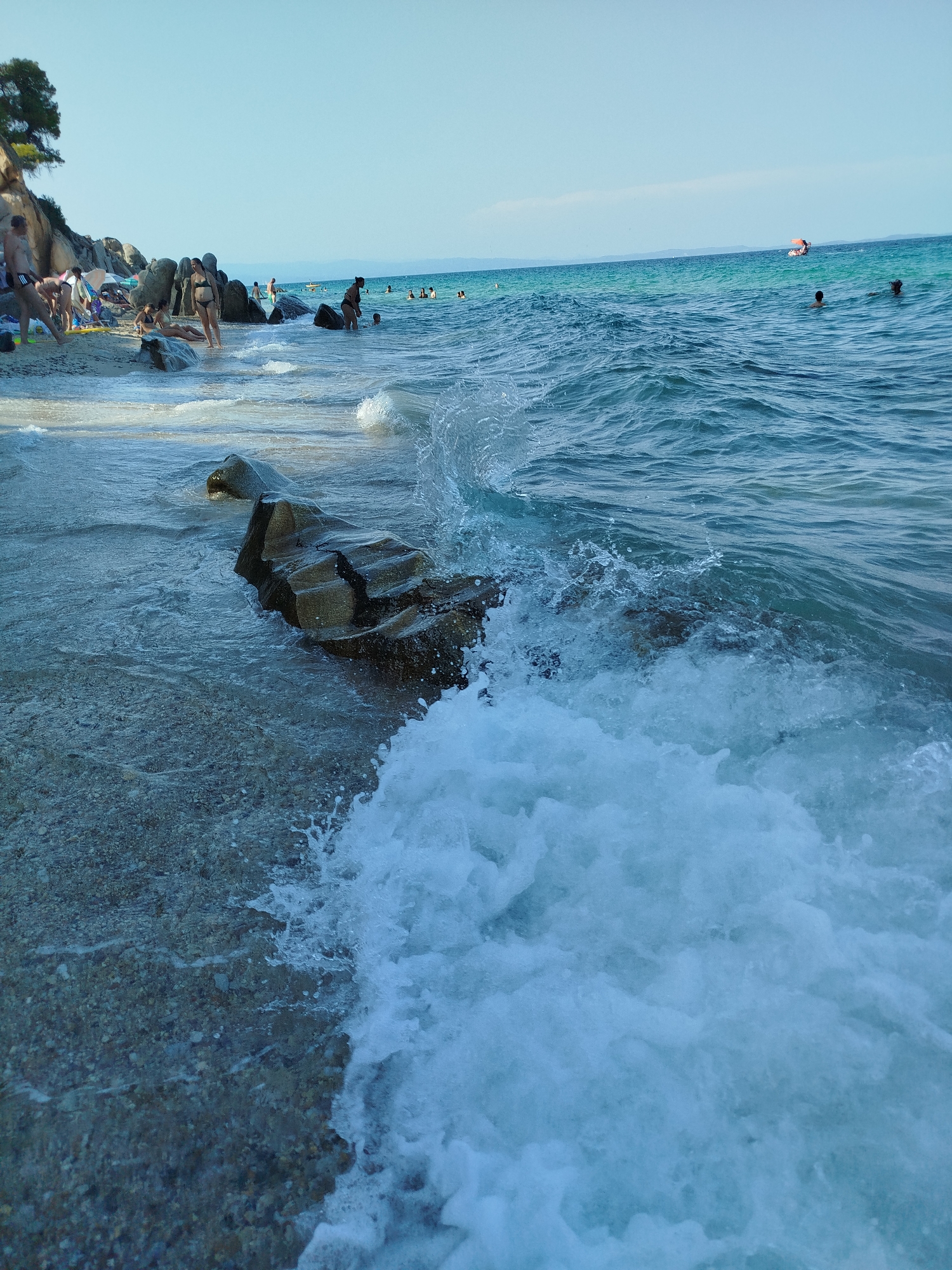 This is a photograph of the beach in Sithonia Municipal Unit, Greece. The image captures a view of the sea from the shoreline. In the foreground, a wave is crashing onto rocks and the pebbly shore. The water is blue and clear, with white foam from the wave. In the mid-ground, several people are visible in the water and on the beach. Some are swimming, while others are relaxing on the sand. In the background, there are more people, rocks, and a tree on a cliff. The sky is clear and blue. The scene depicts a typical summer day at a Greek beach.