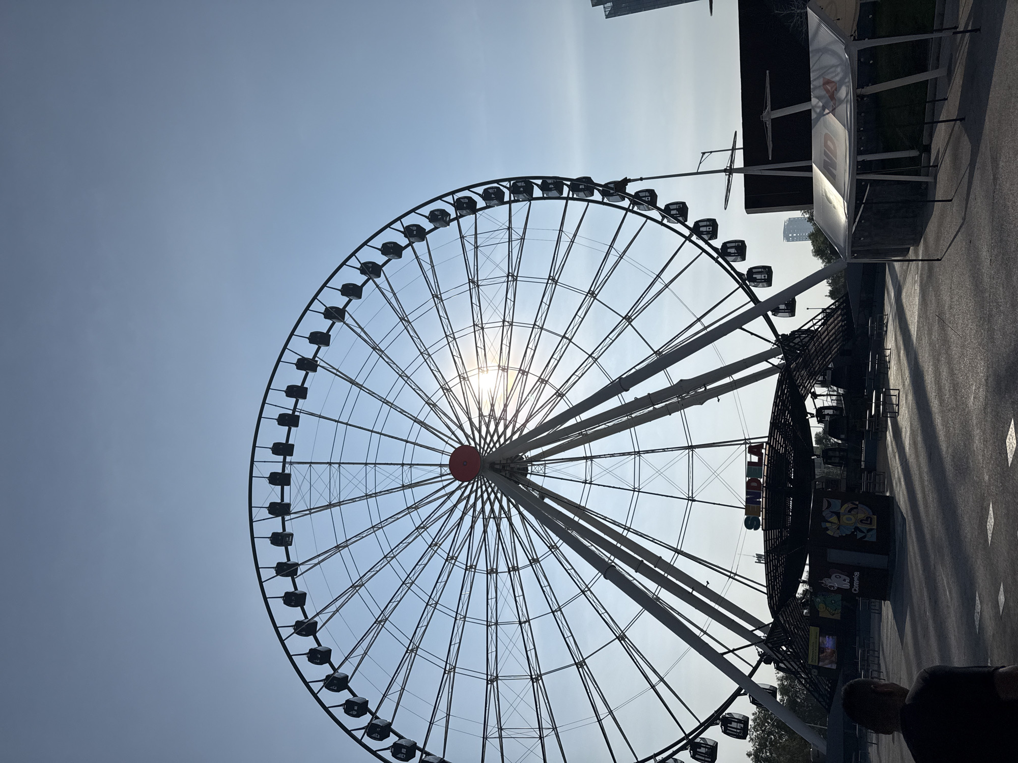 The image depicts La Estrella de Puebla, a Ferris wheel located in Puebla, Mexico. The Ferris wheel is the central subject, dominating the frame with its large circular structure. The sun is positioned directly behind the center of the wheel, causing a starburst effect. The wheel's black gondolas are visible around its circumference. A building and other structures are on the right side of the image. The sky is a pale blue.
