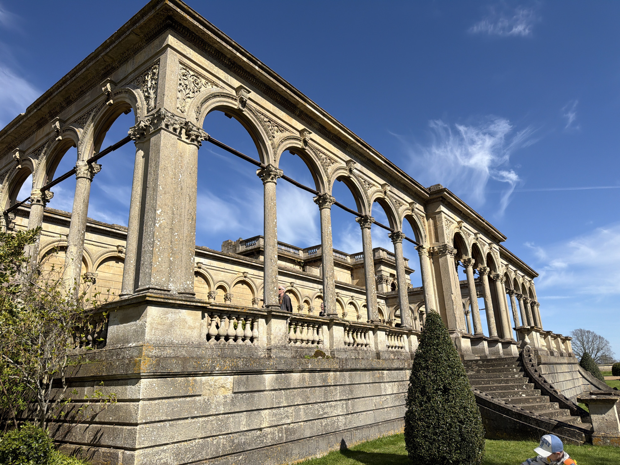 The image depicts an architectural structure in Malvern Hills, United Kingdom. The structure features a series of arches supported by pillars, with intricate decorative details. A balustrade runs along the middle level. Stairs lead up to the structure on the right side. The sky is blue with wispy clouds. The scene is set in a grassy area with a few trees and shrubs, and one person is visible at the bottom right corner of the frame.