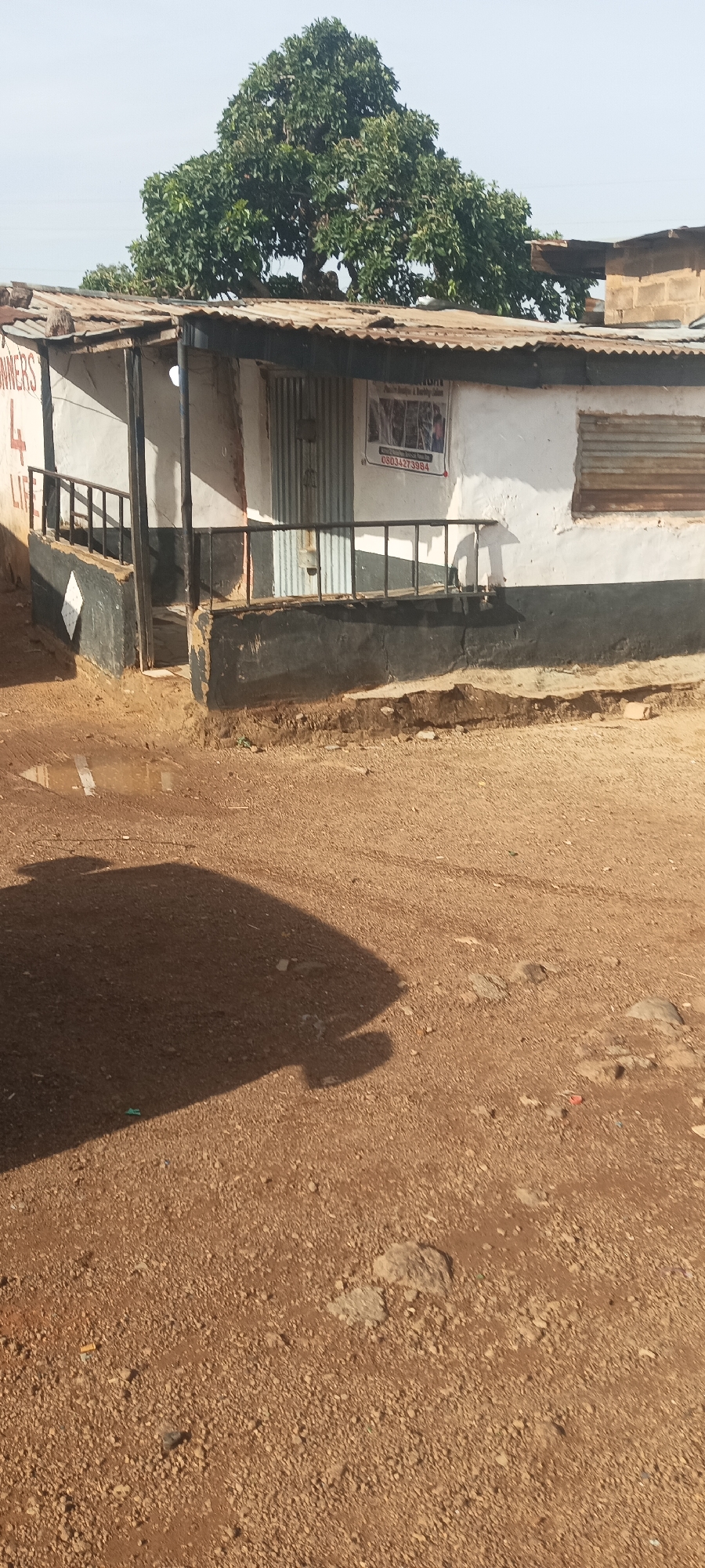 The image shows a weathered, single-story building in Ladura Foron, Nigeria. The building is primarily white with a dark base and a corrugated metal roof. The facade has a small porch with a dark railing. A metal door is visible, and a poster is affixed to the wall near the door. A tree is visible behind the building. The ground in front is a dirt road with a shadow cast in the foreground. There are no people visible in the image.