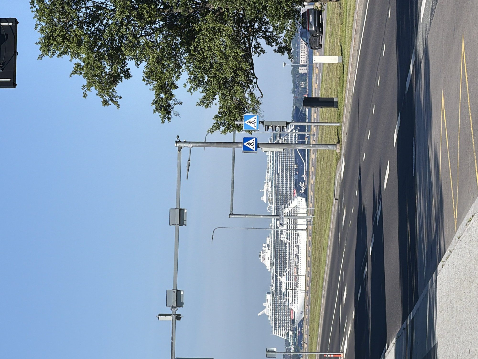 The image captures a wide-angle view of a road in Tallinn, Estonia, with a large cruise ship docked in the background. The scene is illuminated by bright sunlight, casting distinct shadows on the asphalt road. The road itself is marked with white dashed lines and a yellow junction box in the foreground. On the right side of the road, a dark grey car is parked on a grassy verge next to a large, leafy tree.
In the mid-ground, a pedestrian crossing is indicated by two blue and white pedestrian crossing signs mounted on a pole. Further back, the impressive white cruise ship dominates the harbor area, its various decks and structures clearly visible against a hazy blue sky. Several smaller vessels are also present in the water near the cruise ship.
The environment suggests a sunny day in a port city, likely a popular tourist area given the presence of a large cruise ship. The overall impression is one of a modern urban landscape meeting the maritime environment.