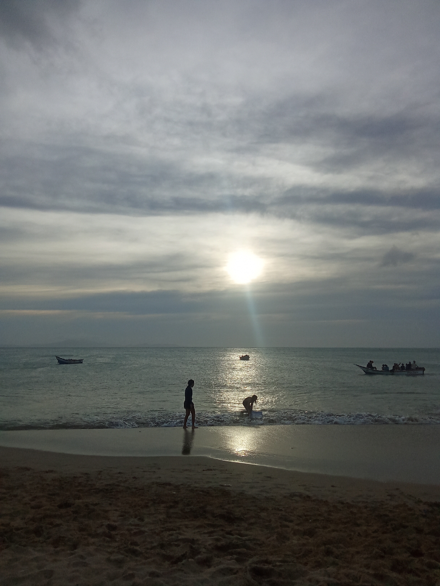 The image depicts a beach scene in Juan Griego, Venezuela. The sky is overcast with the sun visible through a break in the clouds. Three boats are visible on the water, one small boat, one larger boat with multiple people in it, and another boat. Two people are in the water near the shoreline. One person is standing near the shore while another is crouched down in the water. The foreground shows the sandy beach.