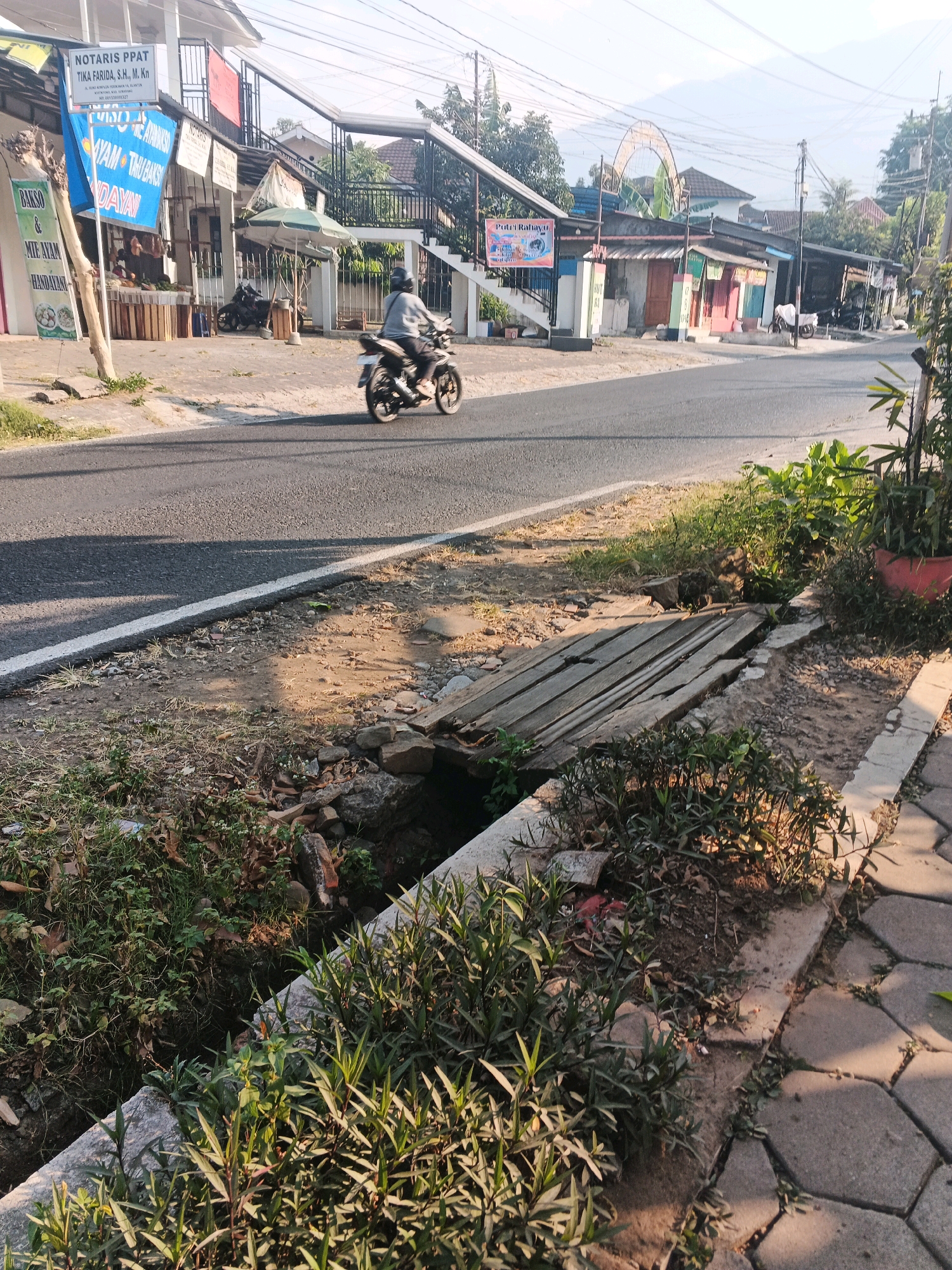 The image captures a street scene in Ungaran, Indonesia, on a sunny day. In the foreground, a narrow, overgrown drainage ditch runs parallel to a paved sidewalk. A makeshift wooden bridge, constructed from planks, spans the ditch. Beyond the ditch and sidewalk, a paved road is visible, with a white line marking the edge. A motorcycle rider, seen from the back, is traveling away from the camera on the road. On the left side of the road, several small shops and businesses are present, indicated by signage. One sign, in blue, reads "AYAM THU BAKSO HINDAYANI," suggesting it's a food stall. Another sign, "NOTARIS PPAT TIKA FARIDA, S.H., M. Kn," identifies a notary office. In the background, buildings with tiled roofs and some vegetation are visible, with a distant, hazy landscape suggesting hills or mountains. The overall impression is of a typical urban street in Indonesia, with a mix of commercial activity and infrastructure elements.