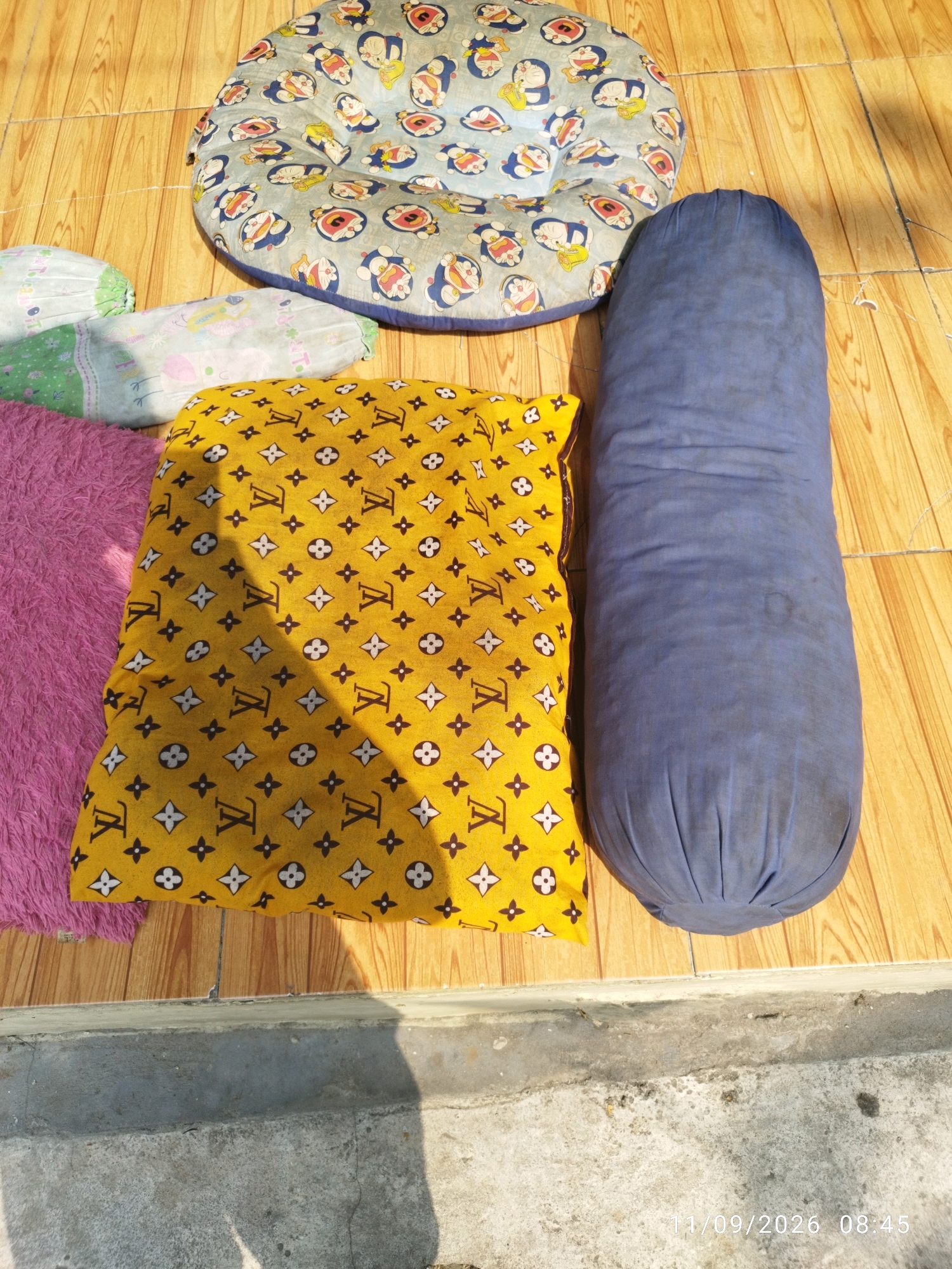 An overhead shot shows several cushions and pillows laid out on a wooden floor. A round, light blue cushion patterned with cartoon characters, possibly Doraemon, is in the upper portion of the image. To the left of this, there are two cylindrical pillows with green and white patterns. Below these and slightly to the right is a yellow pillow with a repeating pattern of what appears to be the Louis Vuitton monogram. To the right of the yellow pillow is a long, dark blue bolster pillow. A small portion of a fuzzy pink mat is visible on the left side of the image. The scene appears to be outdoors, indicated by the shadows cast on the wooden floor and a concrete surface with shadows in the foreground, suggesting it is a sunny day. The timestamp "11/09/2026 08:45" is present in the lower right corner. This collection of soft furnishings is situated on a tiled wooden floor in an outdoor area, possibly a patio or veranda, in Mojokerto, Indonesia.