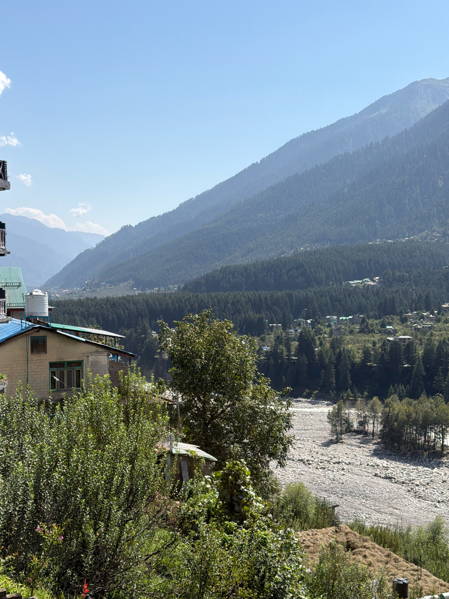 The image depicts a scenic view of a mountain valley in Vashisht, India. In the foreground, a building with a light-colored brick facade and a blue roof is partially visible, adorned with a large white water tank on its roof. Dense green foliage surrounds the building, with some trees reaching towards the sky.
Beyond the building, the landscape opens up to a wide, rocky riverbed, suggesting a dry or low-flow season. A cluster of trees stands on the opposite bank of the river, casting shadows on the stony terrain.
The mid-ground is characterized by a lush forest of coniferous trees that blankets the gentle slopes of the mountains. Scattered amongst the trees are small settlements, indicated by buildings with visible roofs.
In the background, majestic mountains rise, their slopes also covered with dense vegetation. The sky is a clear, bright blue, with a few wispy clouds present, suggesting a pleasant day. The overall impression is one of natural beauty and rural tranquility.
