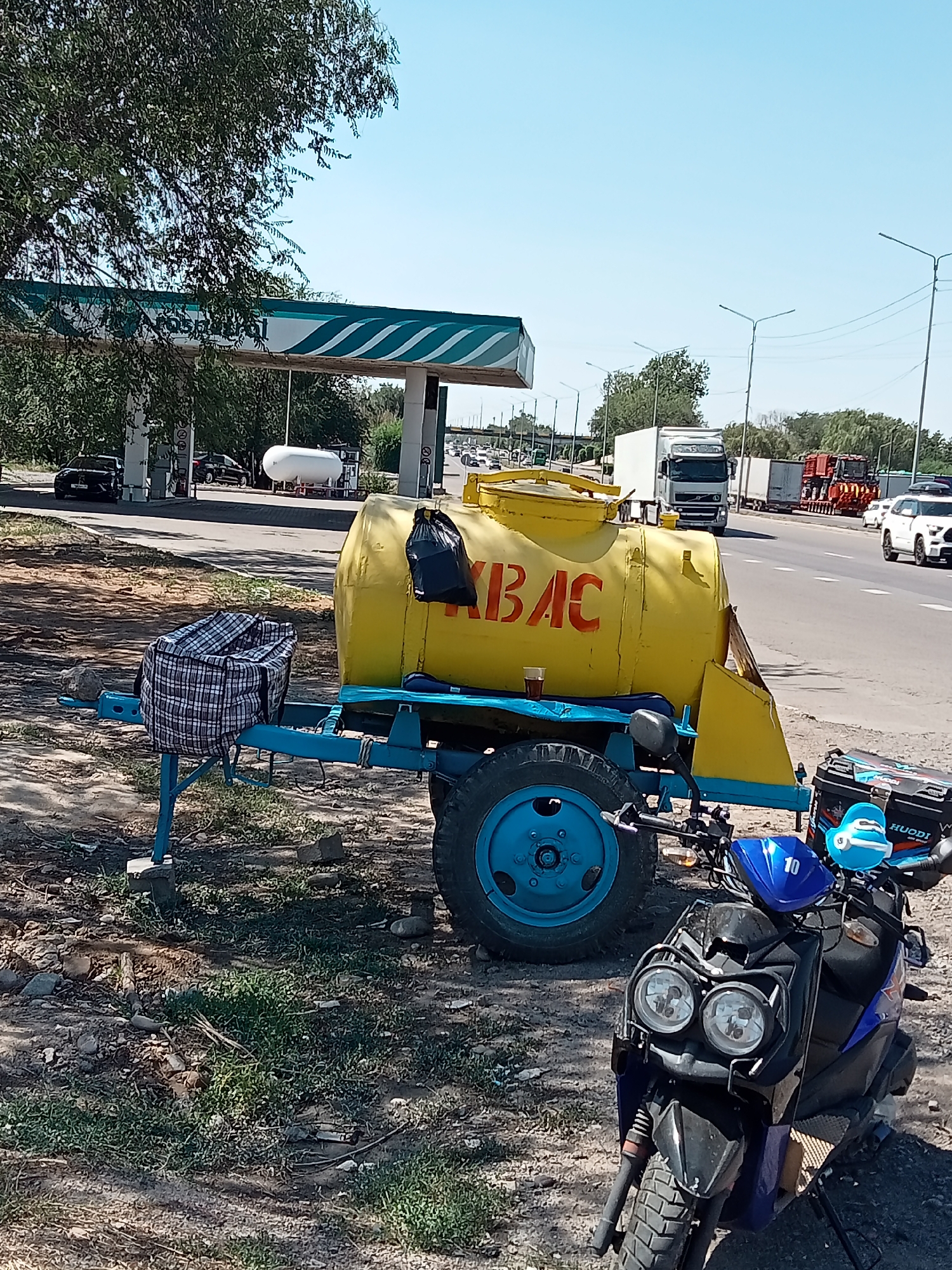 The image shows a scene in Qaskelen, Kazakhstan, on a sunny day. In the foreground, a blue-wheeled trailer with a large yellow tank is parked. The tank has red Cyrillic lettering that appears to spell "КВАС," which refers to a fermented beverage. A black plastic bag is tied to the top of the tank. A plaid bag rests on the trailer's frame. A blue scooter is parked to the right of the trailer, with a blue duck-shaped accessory attached to its handlebars. In the background, a gas station with a white and green striped canopy is visible. Several cars and trucks are on a highway. The environment is dry with sparse vegetation and some loose stones on the ground. The sky is clear and blue.