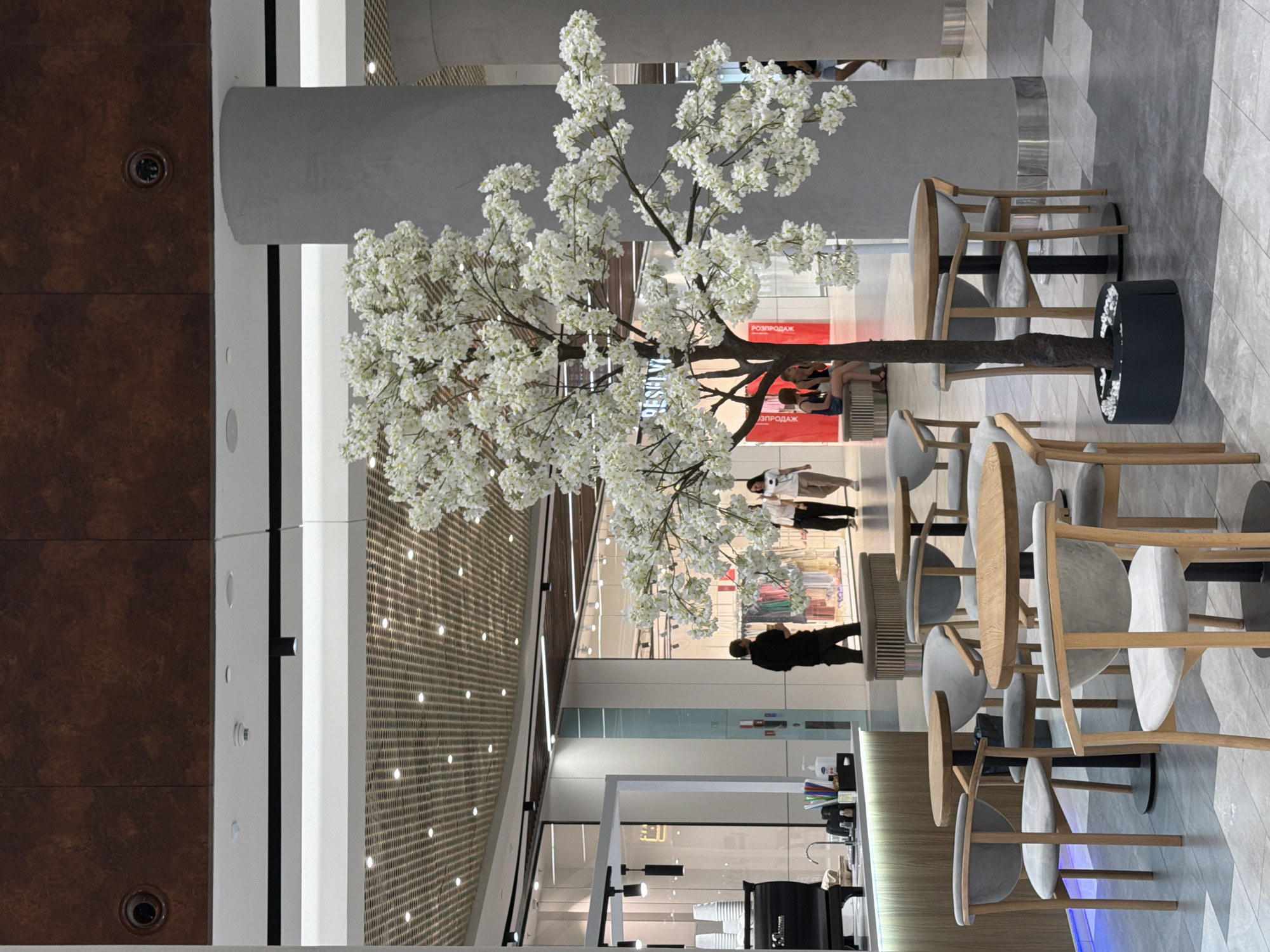 The image depicts an interior scene, likely a shopping mall or a cafe within a mall. In the foreground, several round wooden tables are paired with grey upholstered chairs. A prominent artificial tree with white blossoms stands centrally, positioned within a dark circular planter.
To the left, a coffee counter is visible with a black espresso machine, stacks of paper cups, and various condiments. In the background, several people are walking and interacting. Some are browsing in shopfronts with clothing displays. A large grey cylindrical column is on the right side of the image. The ceiling features a grid-like structure with embedded lights. The floor is tiled with a geometric pattern.
Based on the available information, this scene is located at the Ocean Plaza shopping mall in Kyiv, Ukraine.